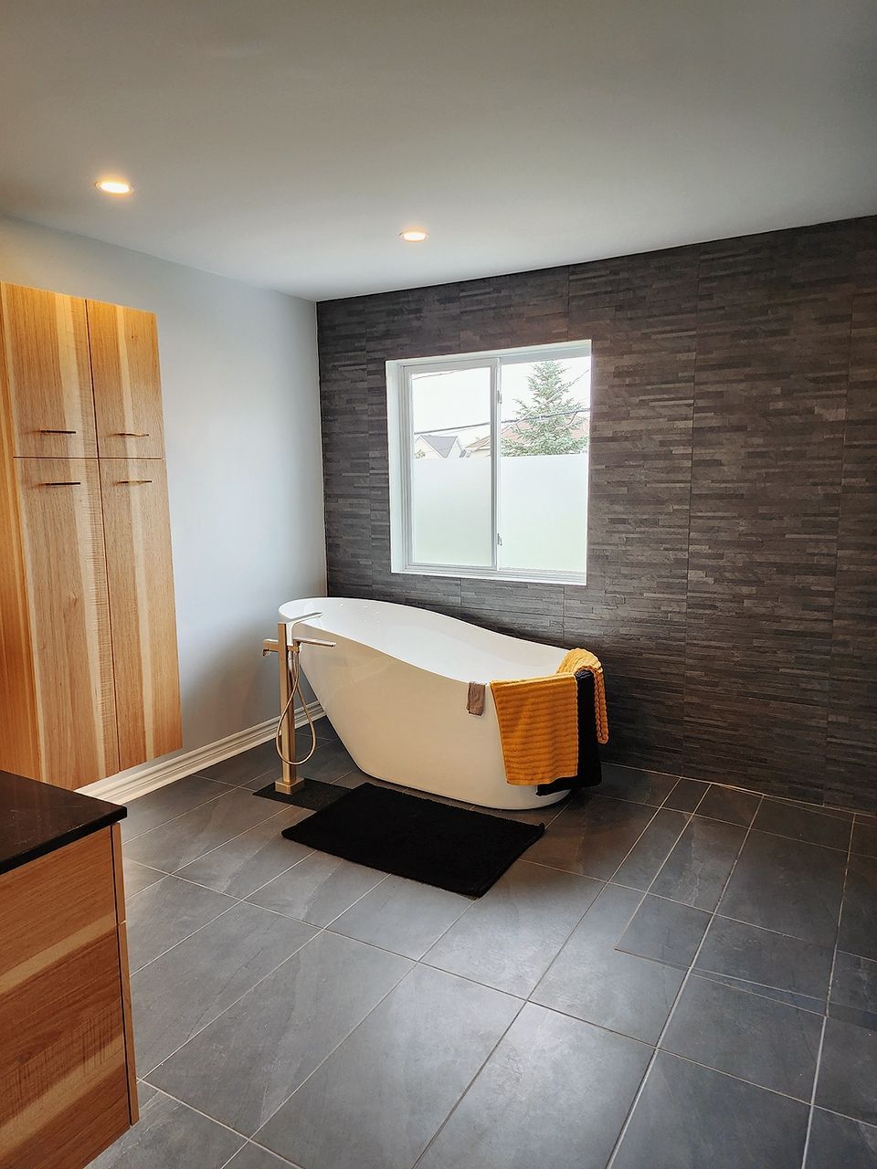 Salle de bain moderne avec baignoire autoportante, mur d'accent en carrelage foncé, armoires en bois et sol en carrelage gris.