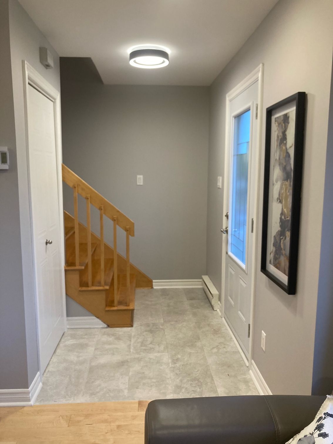 Couloir avec escalier, porte d'entrée et œuvre d'art encadrée. Murs gris clair, sol gris, escalier en bois.