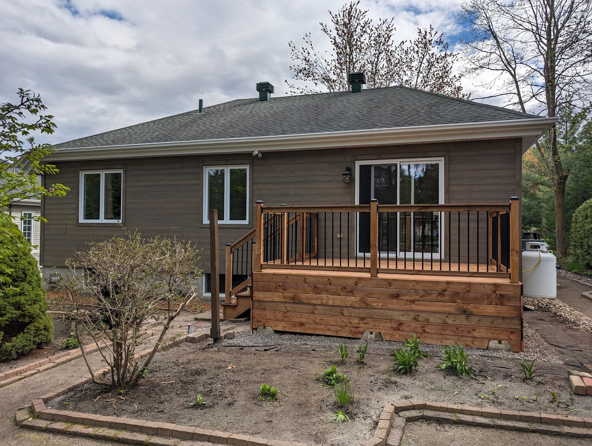 Maison aux façades brunes avec terrasse en bois neuve et porte coulissante en verre. Ciel nuageux, jardin paysager.