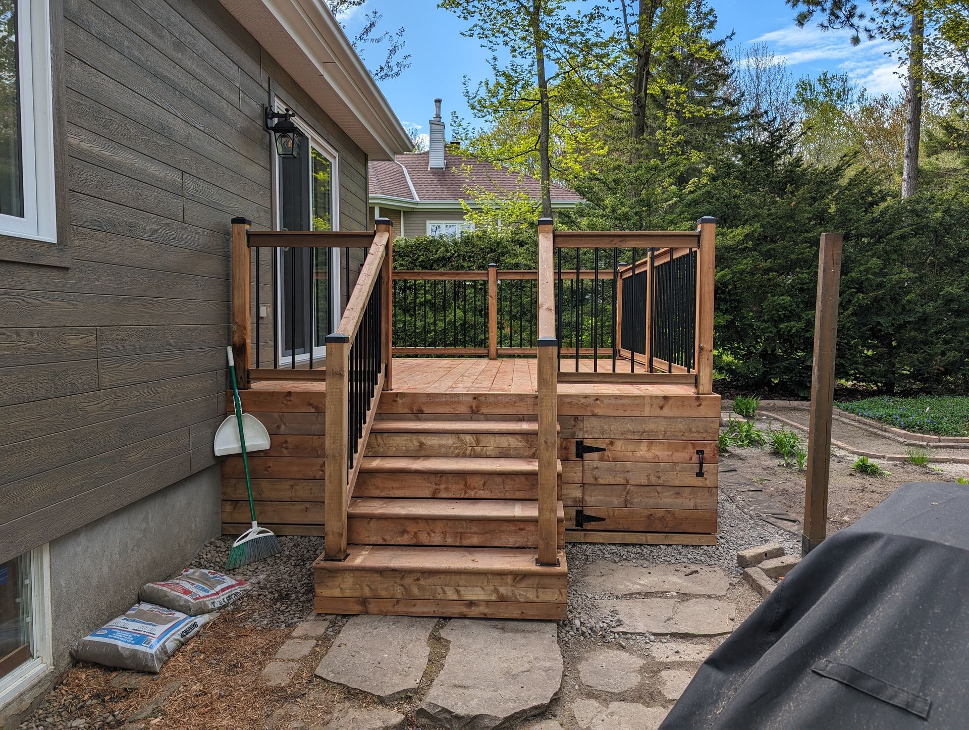 Terrasse en bois avec escalier menant à une plate-forme surélevée; balustrade noire, chemin en pierre et extérieur de la maison.