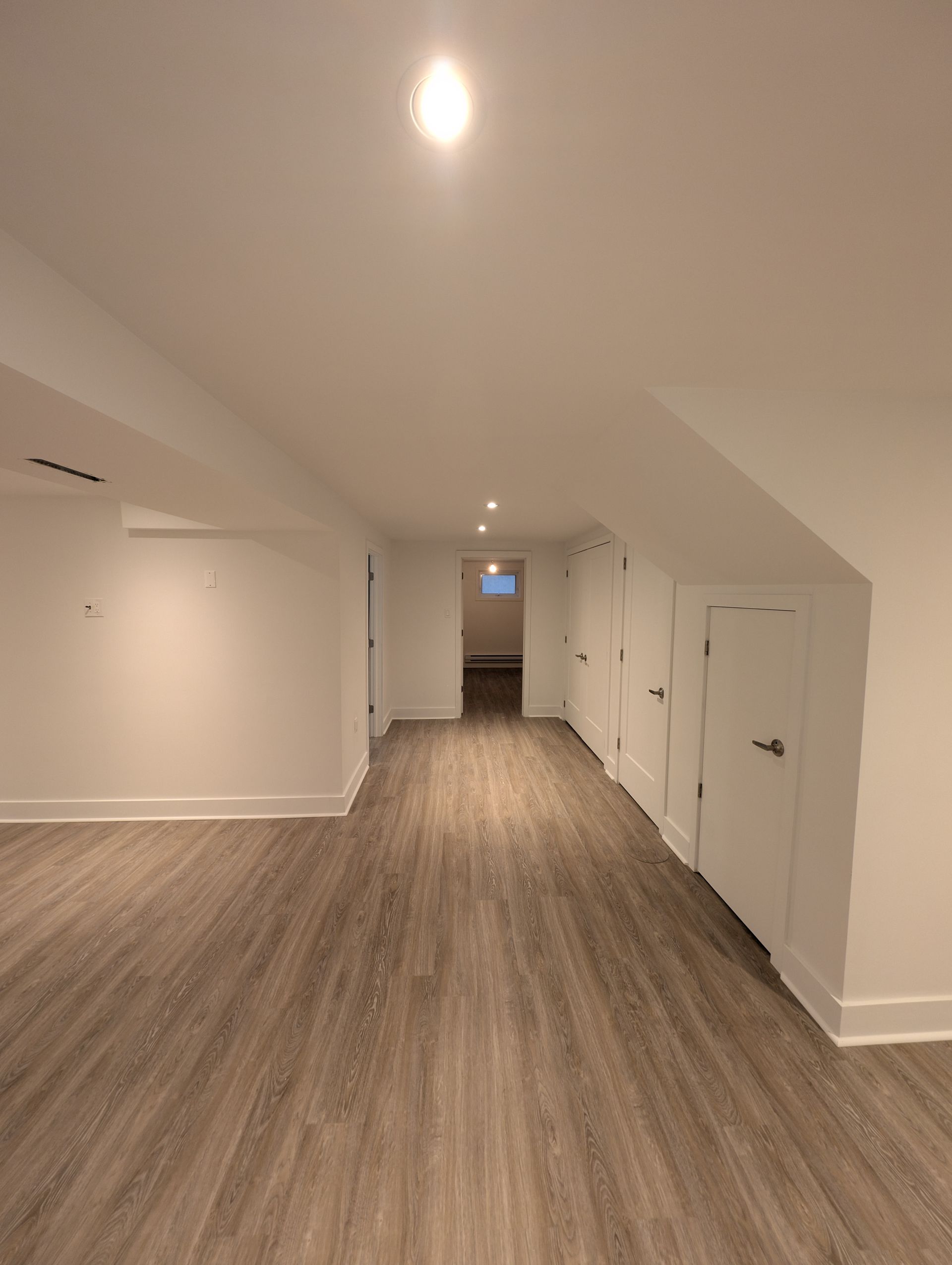 Sous-sol vide aux murs blancs avec plancher imitation bois, placards de rangement et une porte menant à une autre pièce.