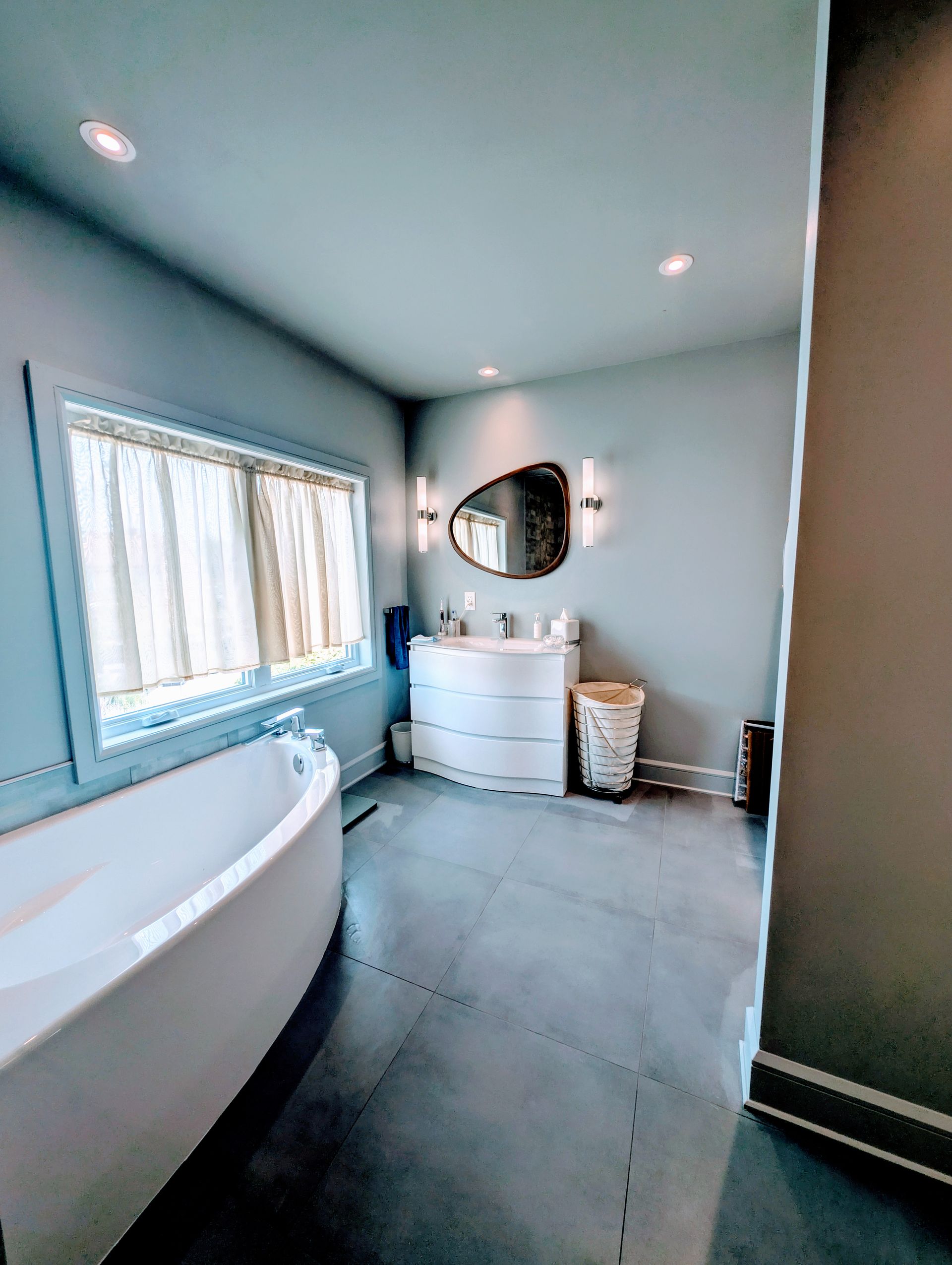 Salle de bain avec grande baignoire, meuble-lavabo, miroir et fenêtre. Murs et sol gris.