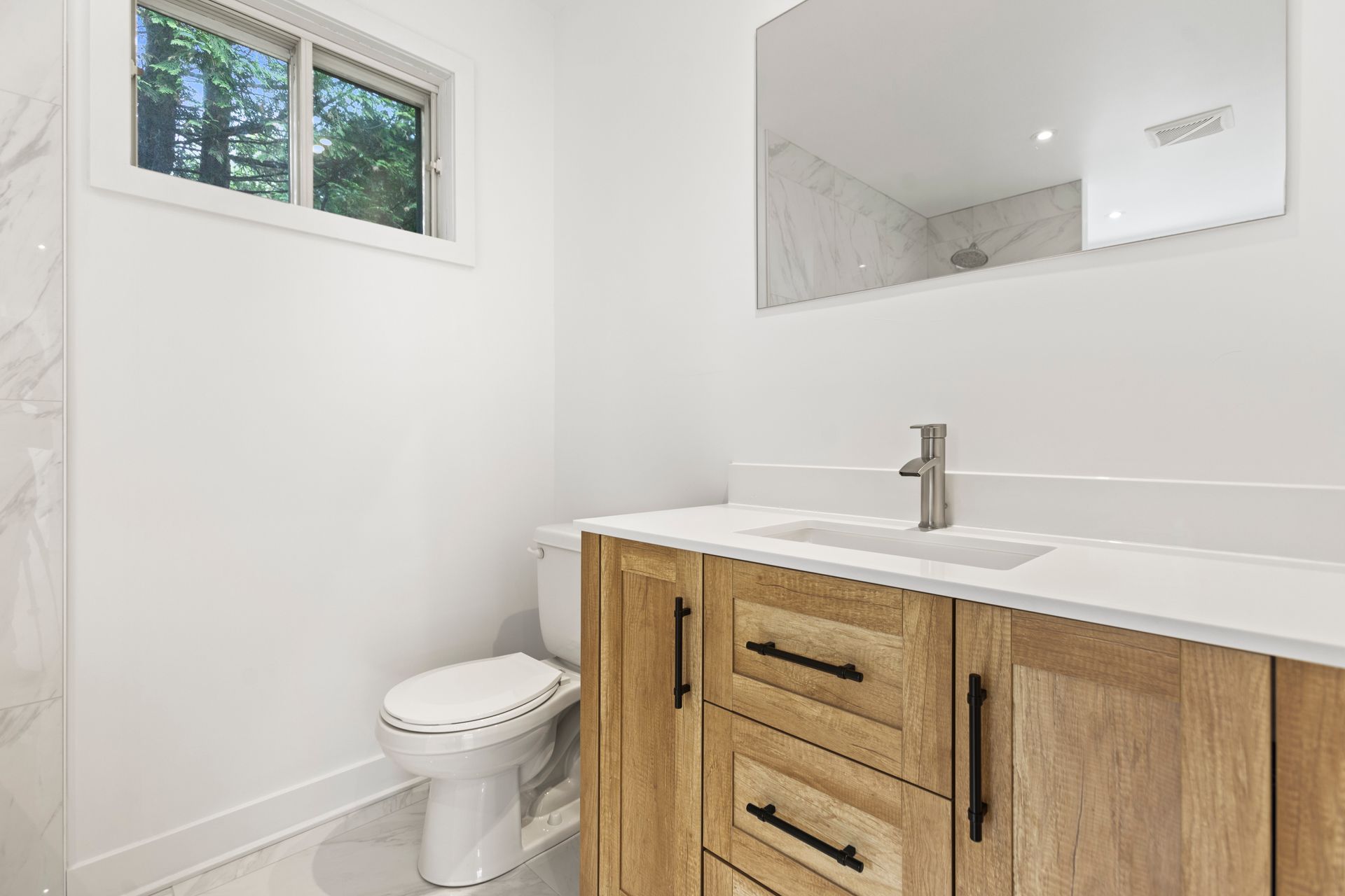 Salle de bain avec meuble-lavabo en bois, plan de travail blanc, toilettes et miroir. Fenêtre à gauche.