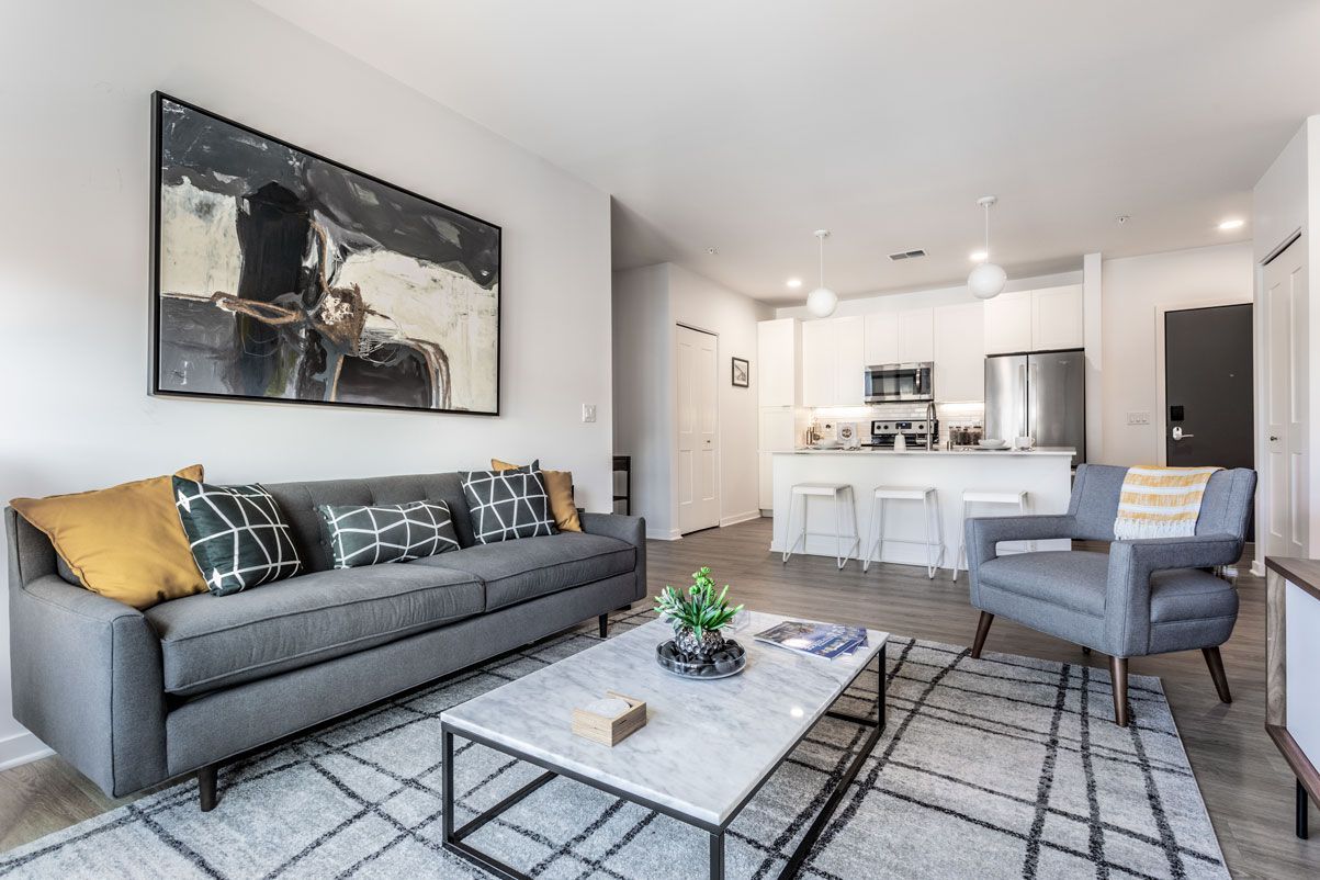 Here at Vim + Vigor Lofts in Wisconsin. A spacious living room features a gray sofa, blue accent chair, modern artwork, warm flooring, and an open kitchen nearby.