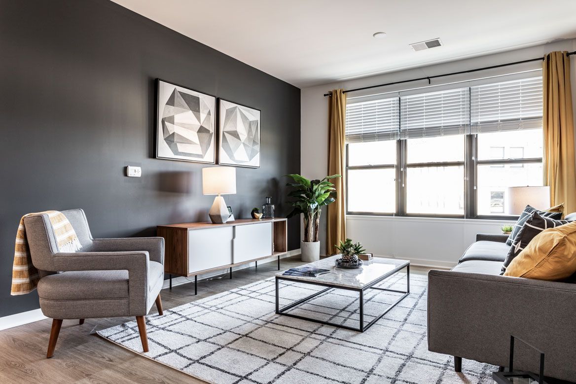 Here at Vim + Vigor Lofts in Wisconsin. A cozy living room features a gray accent chair, gray sofa, patterned rug, warm yellow accents, and large bright windows.