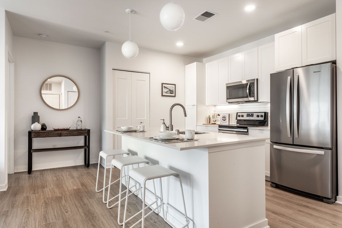 Here at Vim + Vigor Lofts in Wisconsin. A modern kitchen features a spacious island with bar seating, white cabinetry, stainless steel appliances, and warm wood-style flooring.