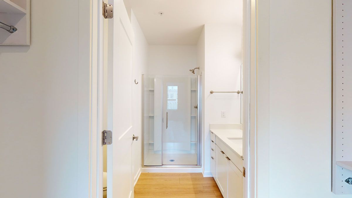 Here at Vim + Vigor Lofts in Wisconsin. A bright hallway view leads to a modern bathroom with a glass shower, white vanity, warm flooring, and nearby closet storage.