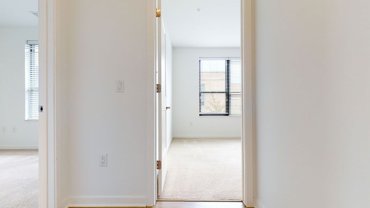 Here at Vim + Vigor Lofts in Wisconsin. A bright hallway view connects two cozy carpeted bedrooms with clean white walls, natural light, and a simple modern layout.
