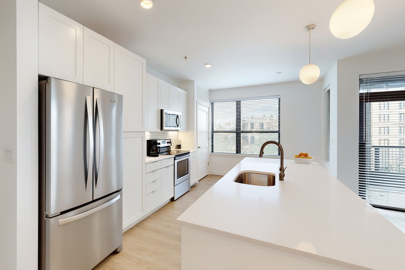Here at Vim + Vigor Lofts in Wisconsin. A modern kitchen features white cabinetry, stainless steel appliances, a spacious island, warm flooring, and bright windows.