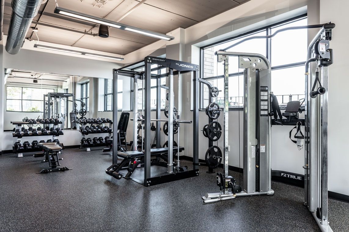 Here at Vim + Vigor Lofts in Wisconsin. A spacious fitness center features strength training equipment, dumbbell racks, large windows, mirrors, and an open industrial-style layout.