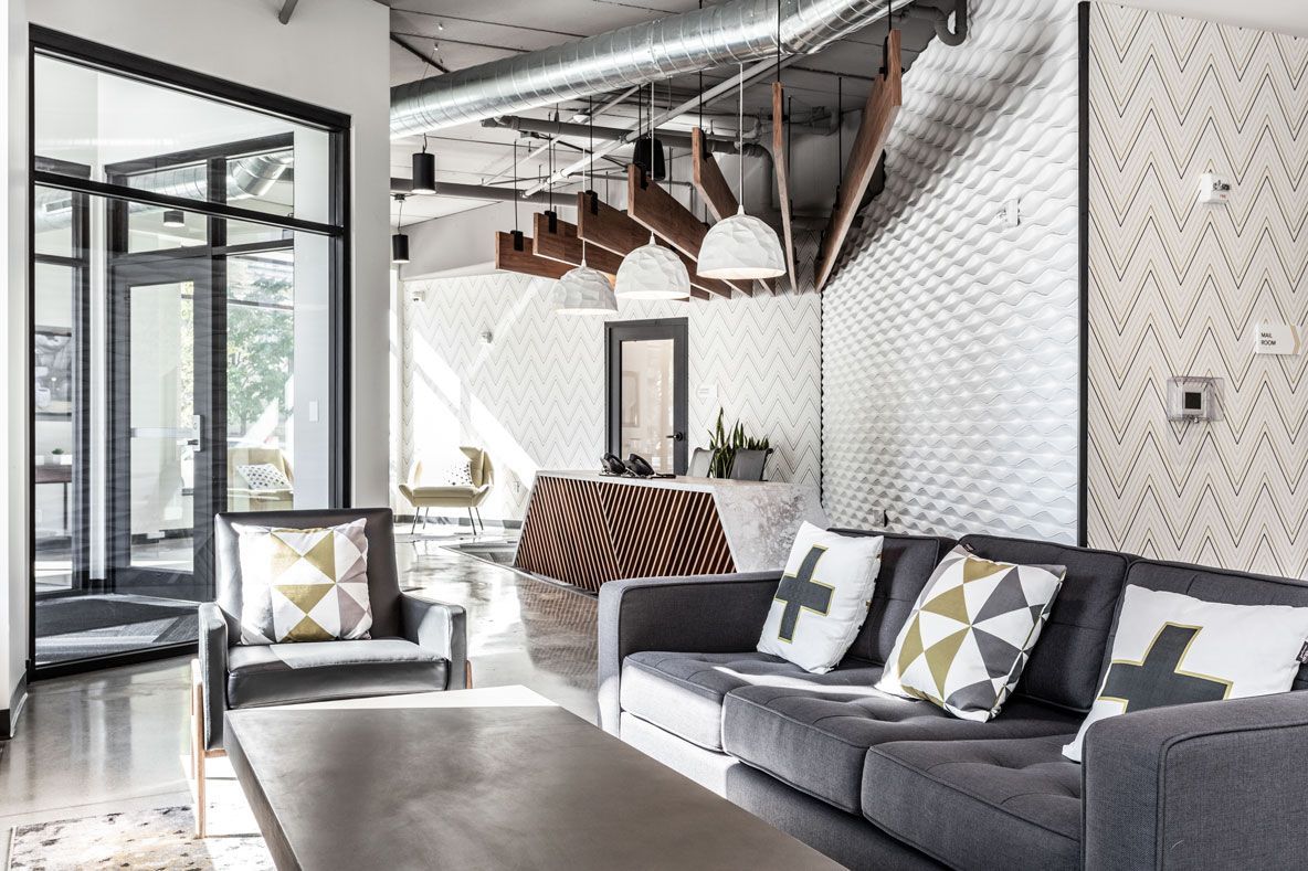 Here at Vim + Vigor Lofts in Wisconsin. A stylish lounge area features a gray sofa, matching accent chair, patterned pillows, modern lighting, and access to the leasing area.