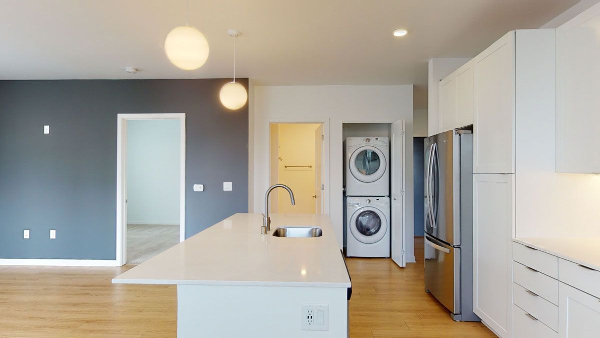 Here at Vim + Vigor Lofts in Wisconsin. A spacious open kitchen features a large white island, stainless steel appliances, in-home laundry, modern lighting, and warm wood-style flooring.