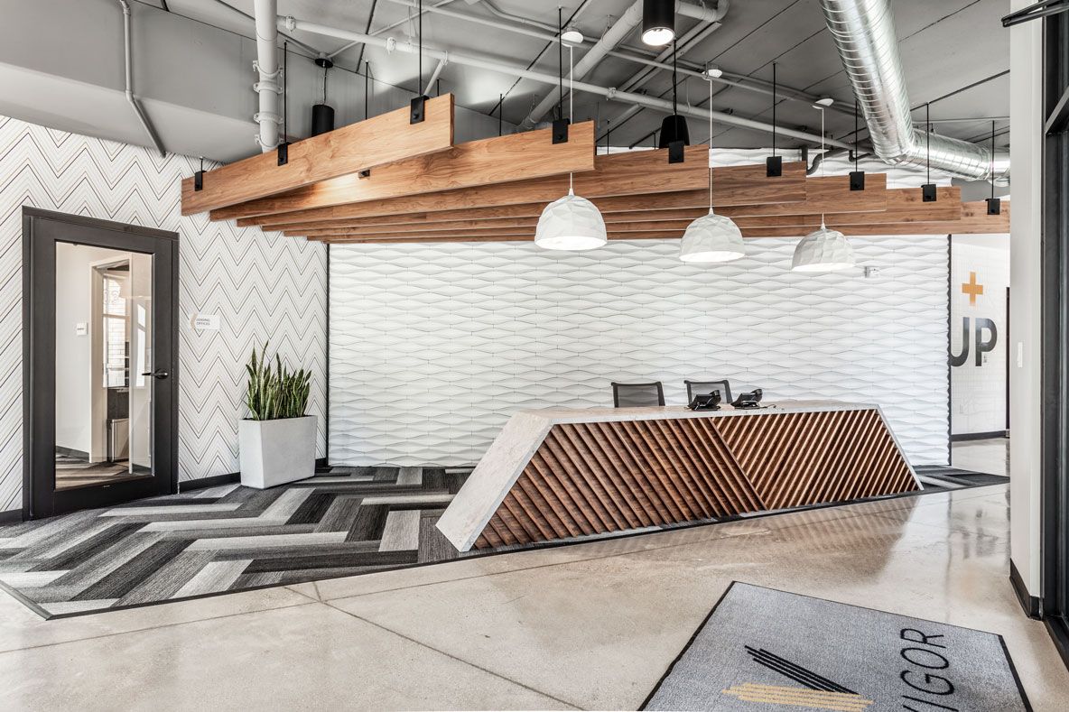 Here at Vim + Vigor Lofts in Wisconsin. A modern leasing lobby features a sculptural front desk, warm wood details, textured white walls, and bright industrial-style lighting.