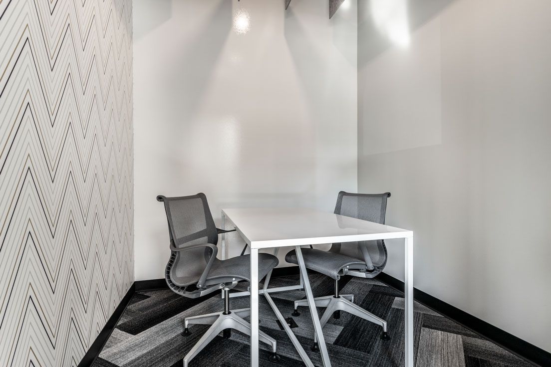 Here at Vim + Vigor Lofts in Wisconsin. A private meeting room features a white table, gray office chairs, patterned carpet, and a clean, modern workspace design.