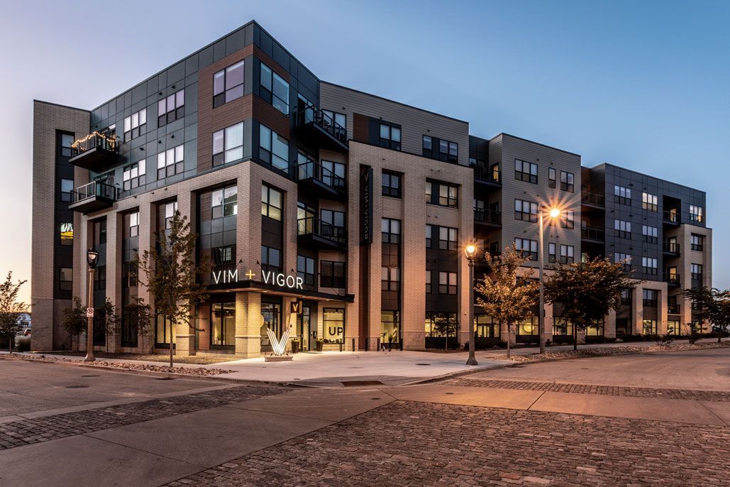 Here at Vim + Vigor Lofts in Wisconsin. A modern exterior view shows the apartment building at dusk with warm lighting, balconies, large windows, and a welcoming entrance.