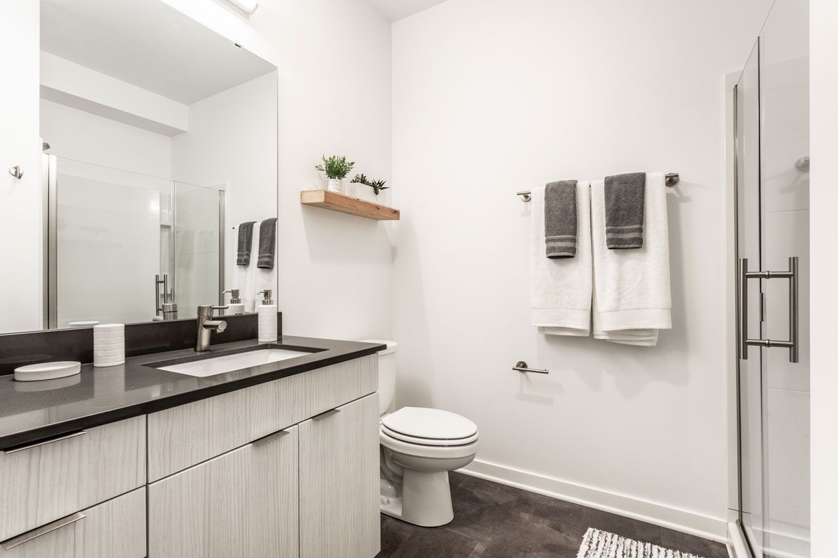 Here at Vim + Vigor Lofts in Wisconsin. A bright bathroom features a modern vanity, glass shower, gray flooring, fresh towels, and simple decorative shelving.