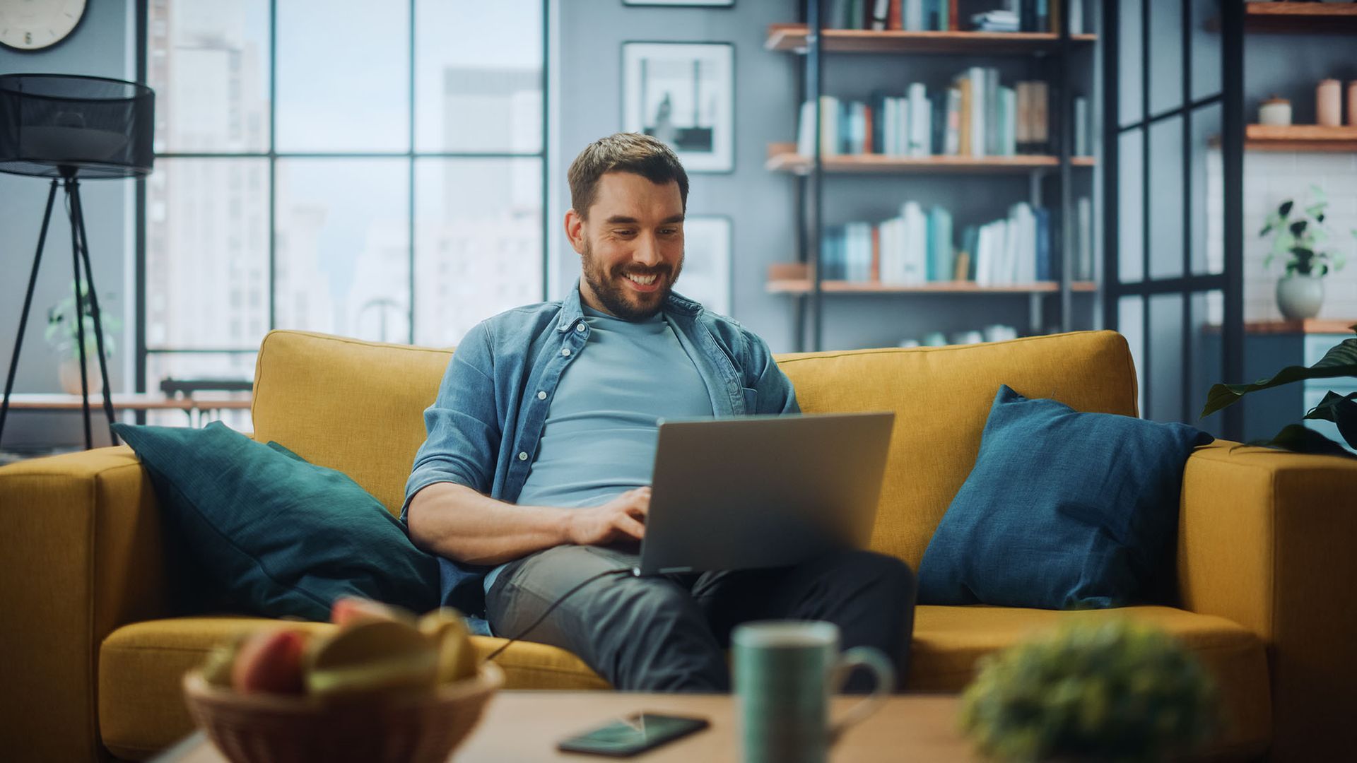 Man on yellow couch working on laptop, smiling. Cozy living room setting.
