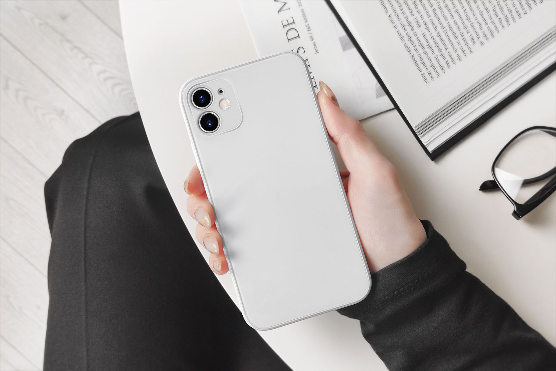 Person holding a white smartphone, sitting near a book and glasses on a white table.