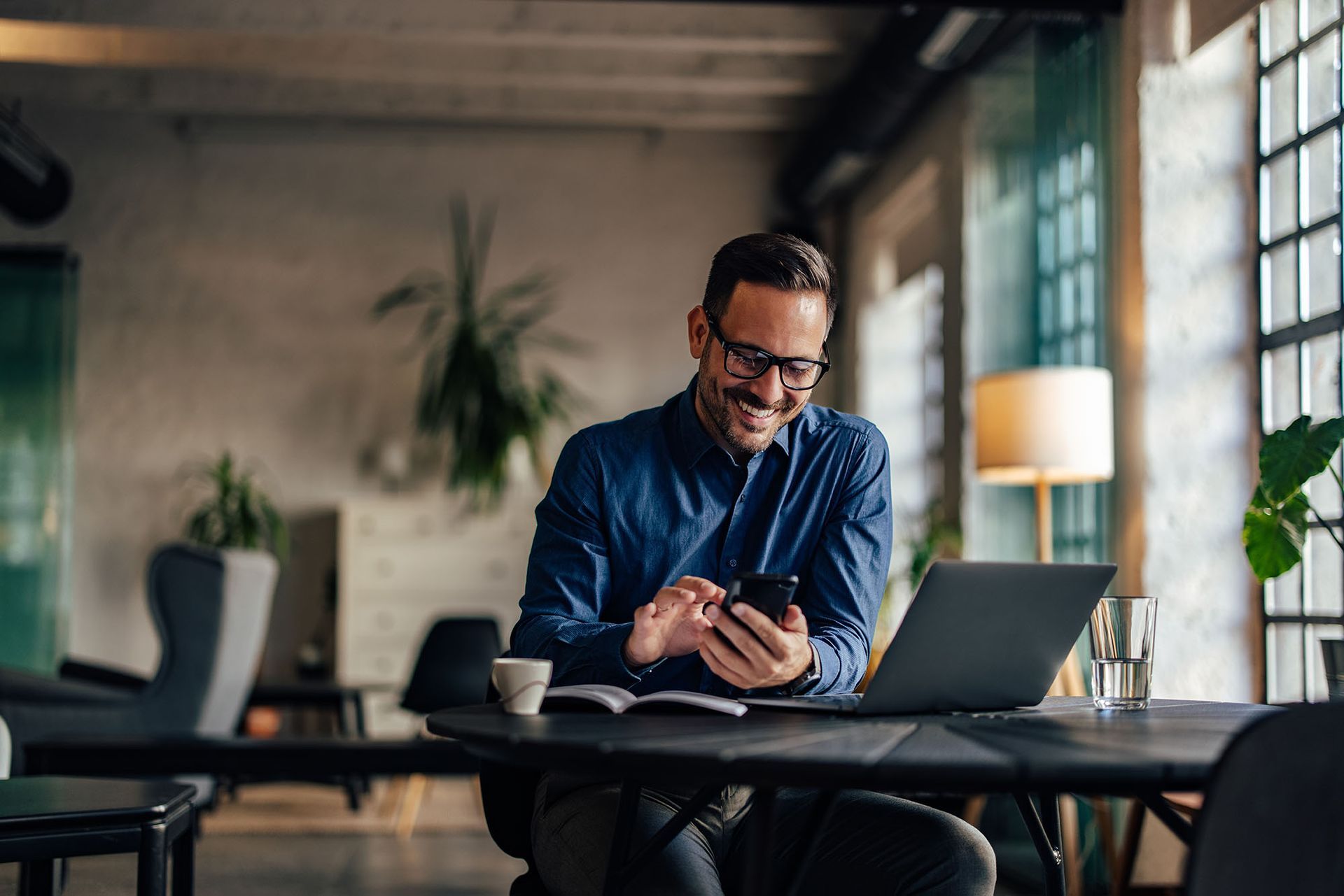 Man wearing glasses smiles while using a smartphone at a desk with a laptop, coffee, and notebook. Man wearing glasses smiles while using a smartphone at a desk with a laptop, coffee, and notebook.