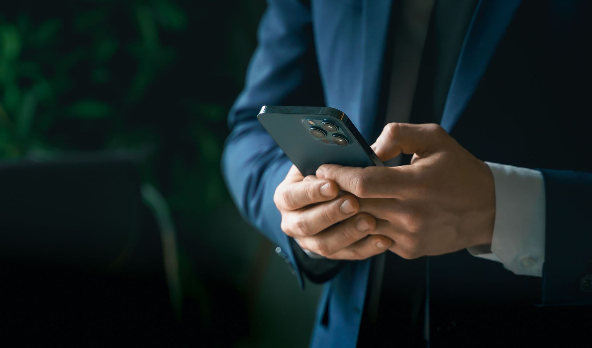 Man in blue suit, hands holding smartphone, typing. Dark background.