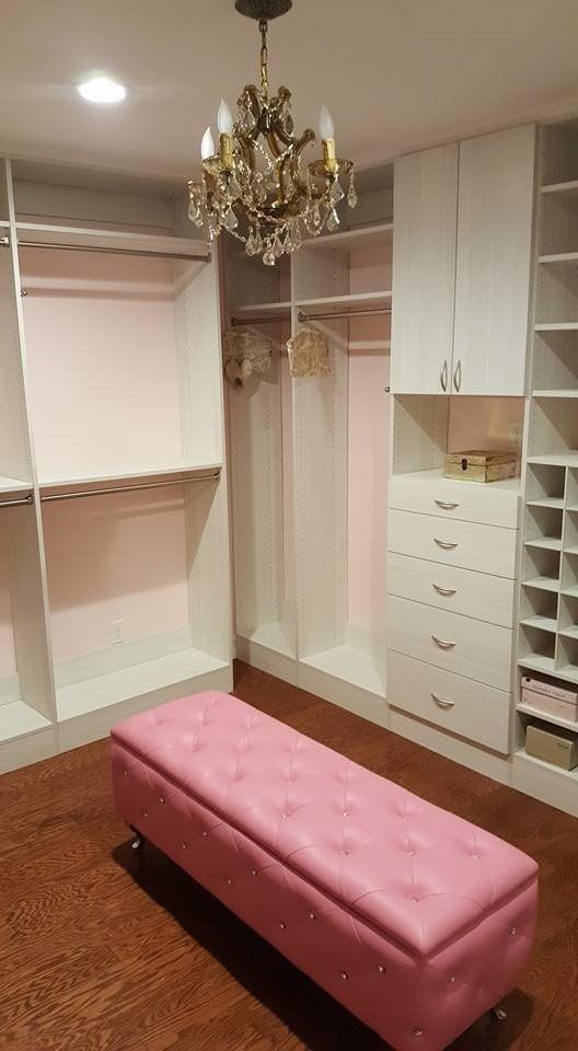 Pink-themed walk-in closet with white cabinets, drawers, and a pink tufted bench under a chandelier.