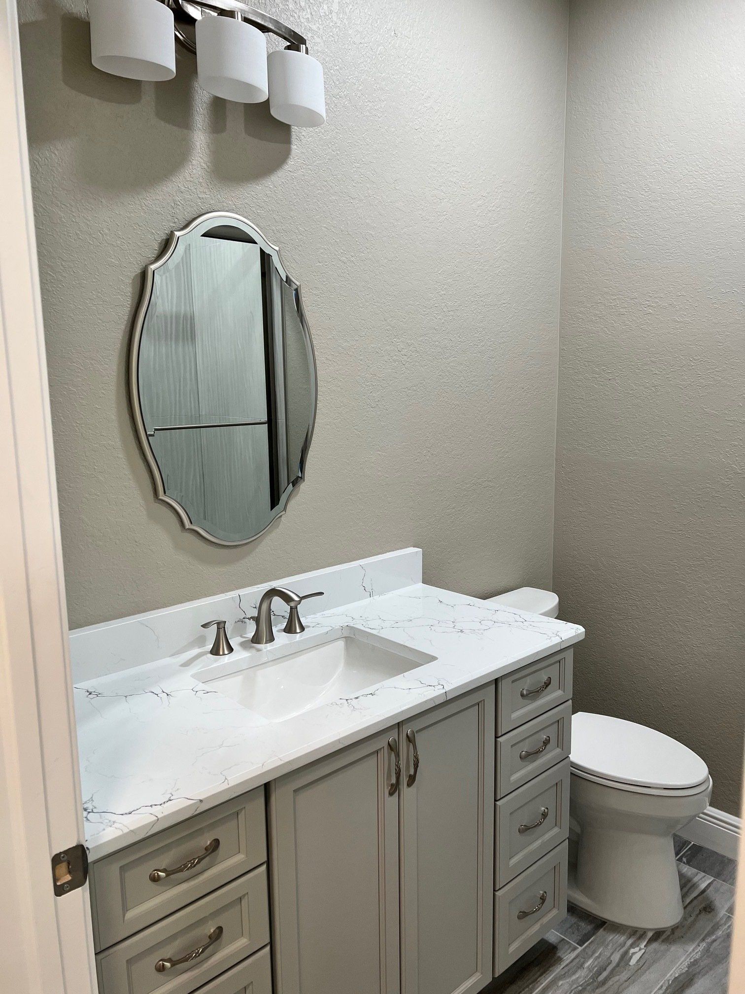 Bathroom with gray vanity, white countertop, and silver framed mirror on sparkly wallpaper.
