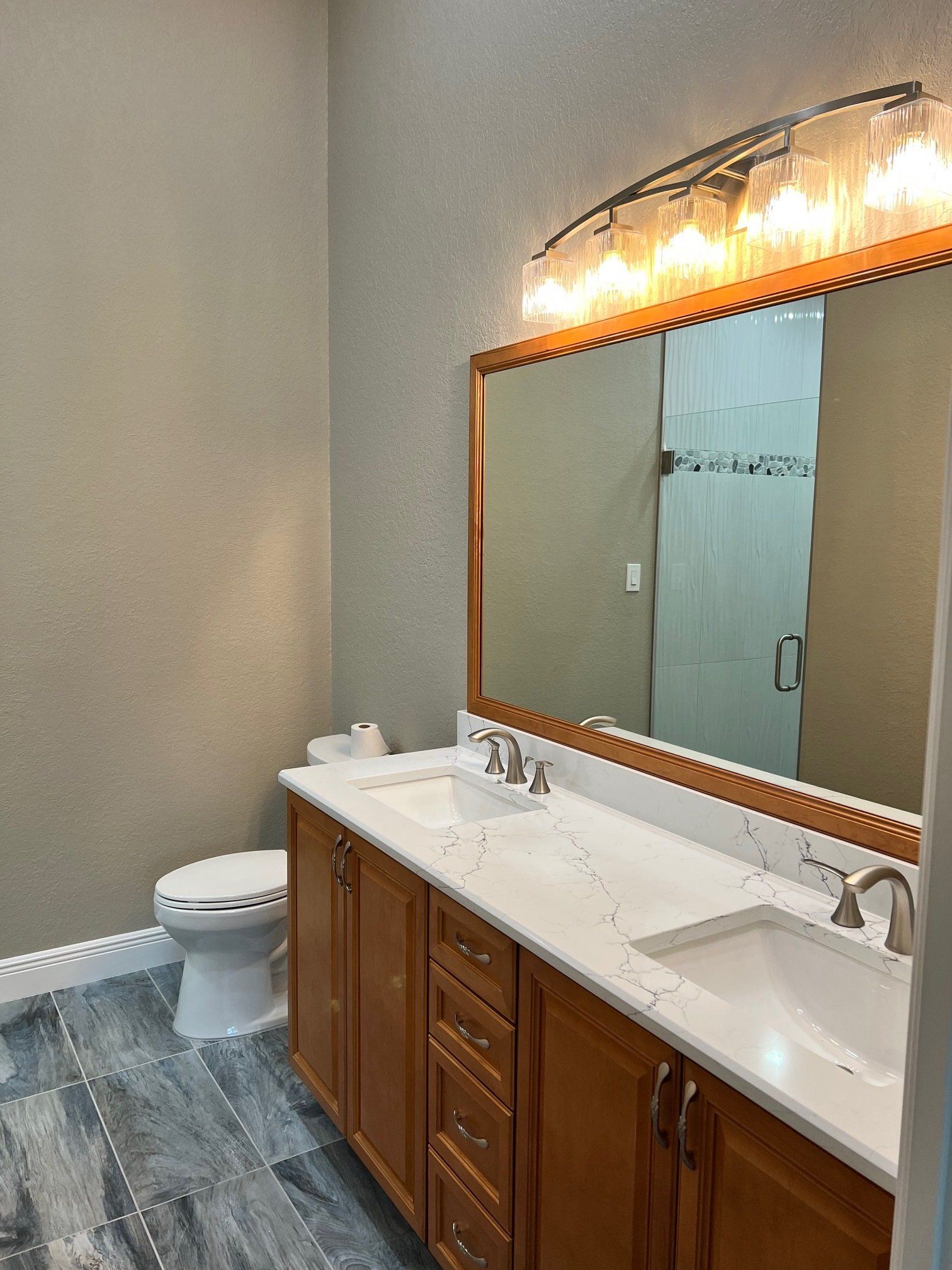 Bathroom with double vanity, toilet, and large mirror. Wooden cabinets, white countertop, and gray tile floor.