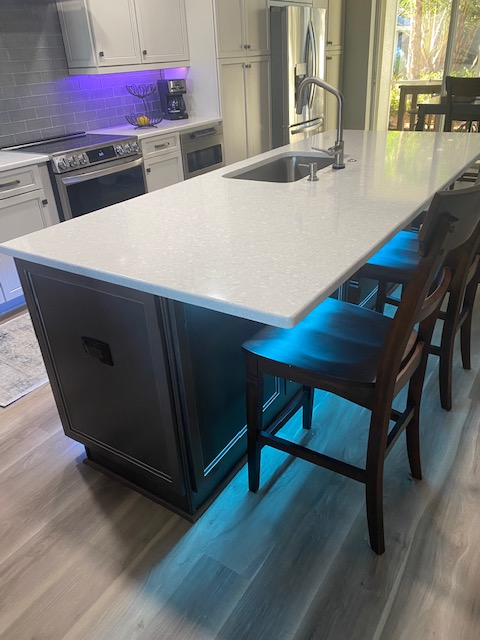 Kitchen island with white countertop, dark cabinetry, seating, and under-cabinet lighting.