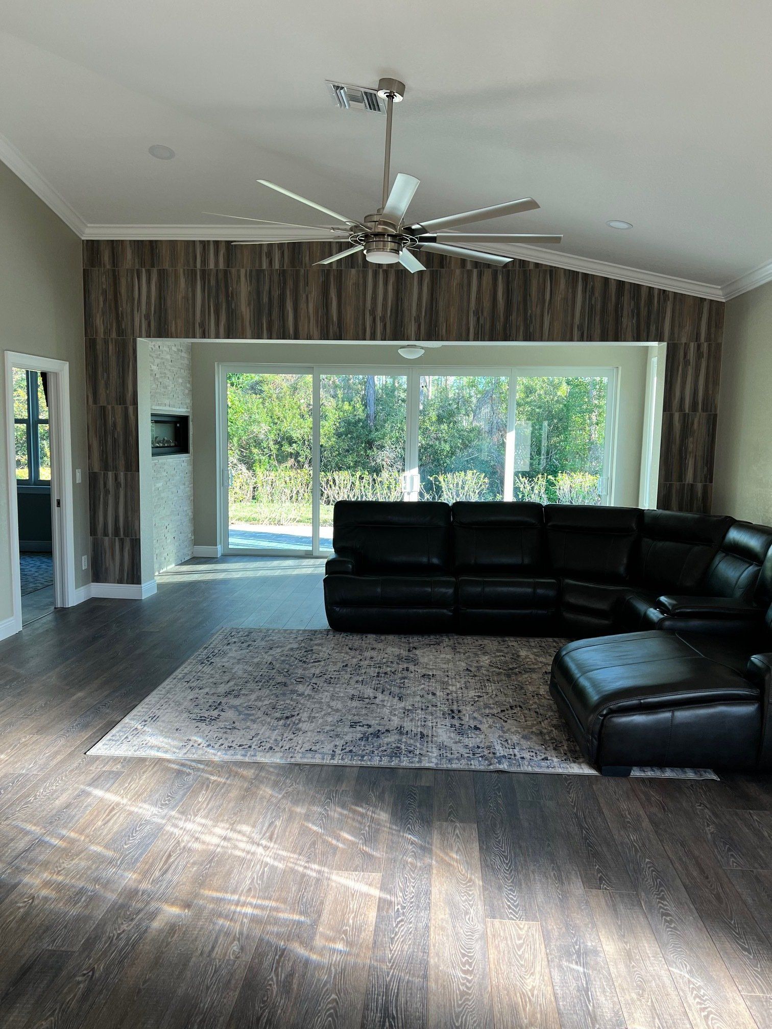 Spacious living room with dark wood flooring, black sectional, and large windows. A ceiling fan and a textured accent wall are present.