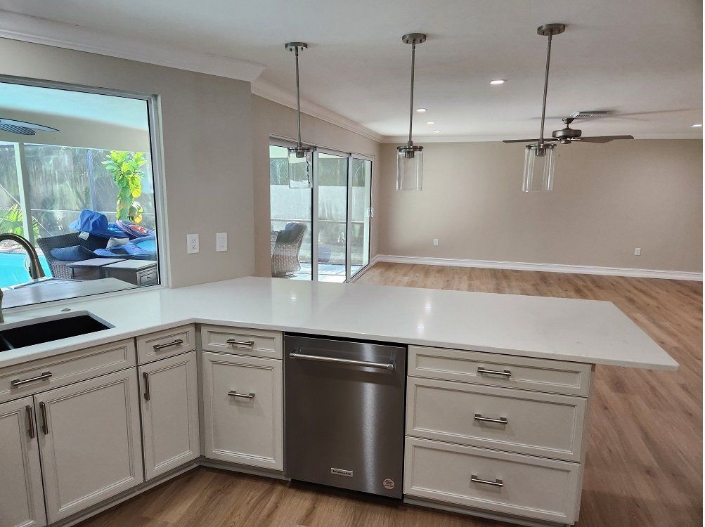 Bright kitchen with white cabinets, island, stainless steel dishwasher, and pendant lights, leading to an open living space.