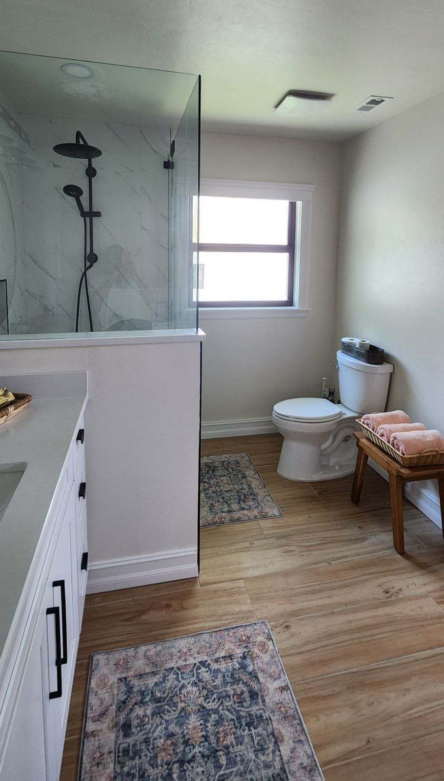 Bathroom with white vanity, glass shower, toilet, bench, and wood-look floor.