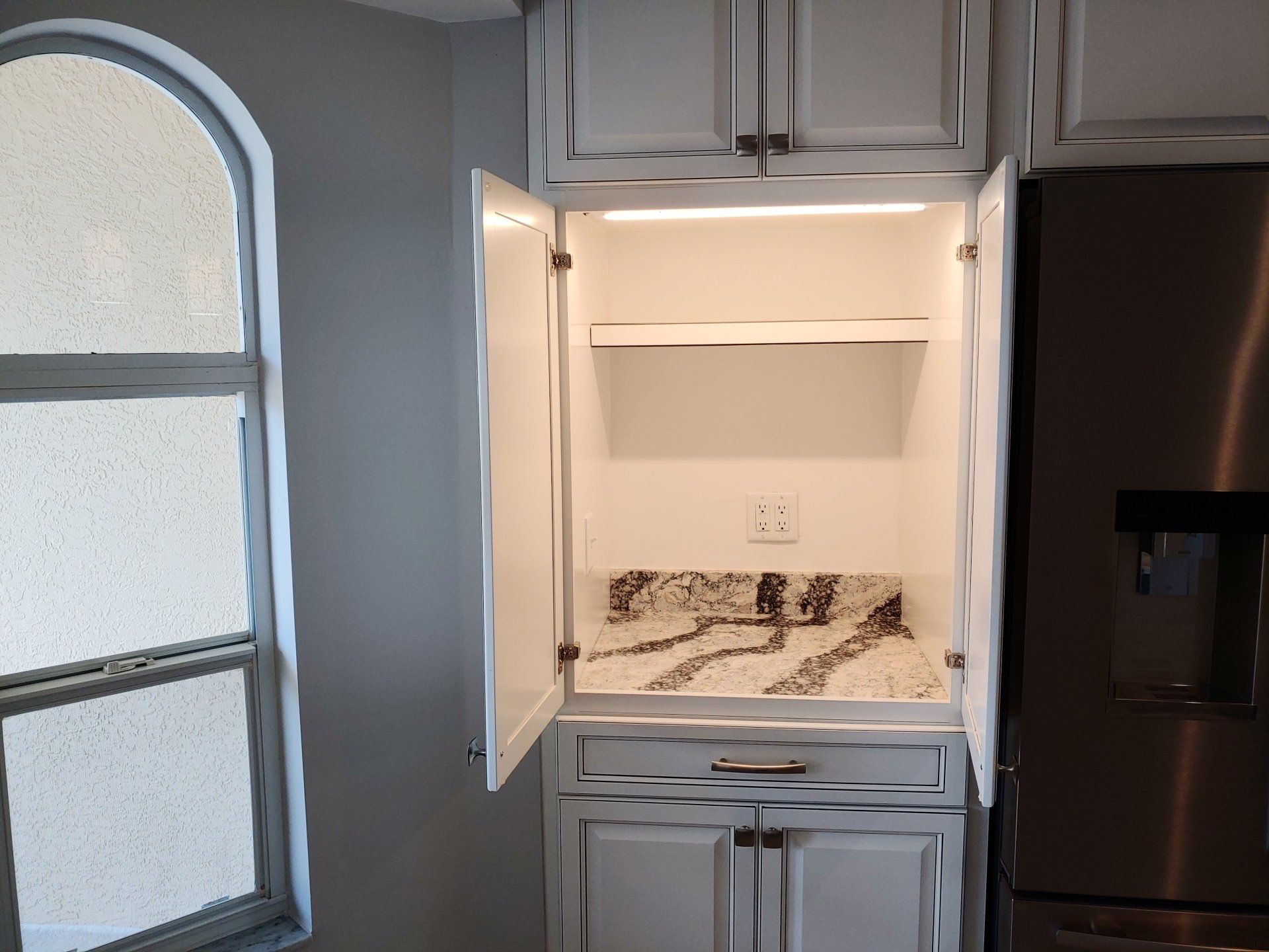 Open white cabinet with shelf, countertop, drawer, and light. Located near a stainless steel refrigerator and a window.