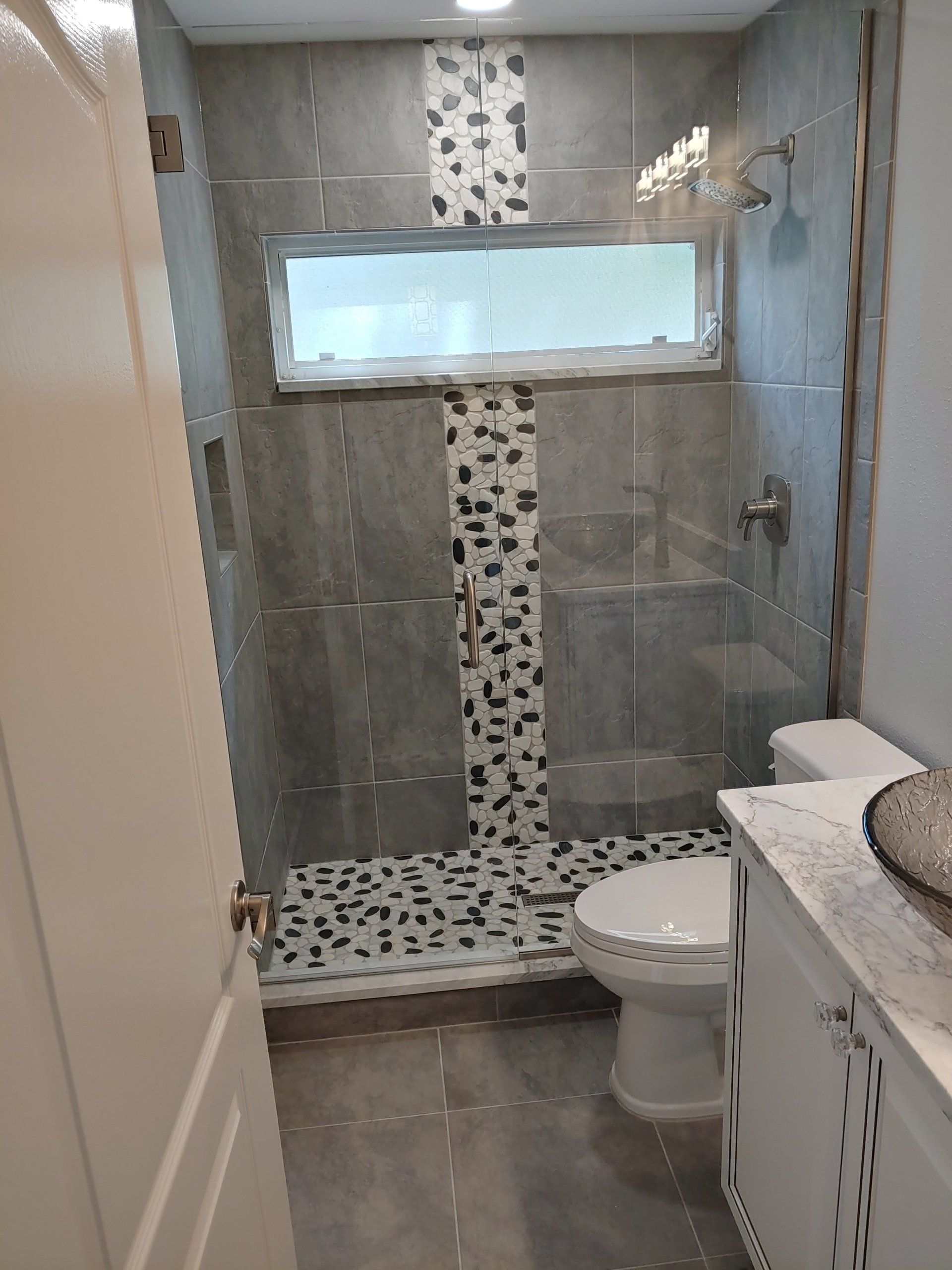 Small gray bathroom with tiled shower, toilet, and vanity; window above.