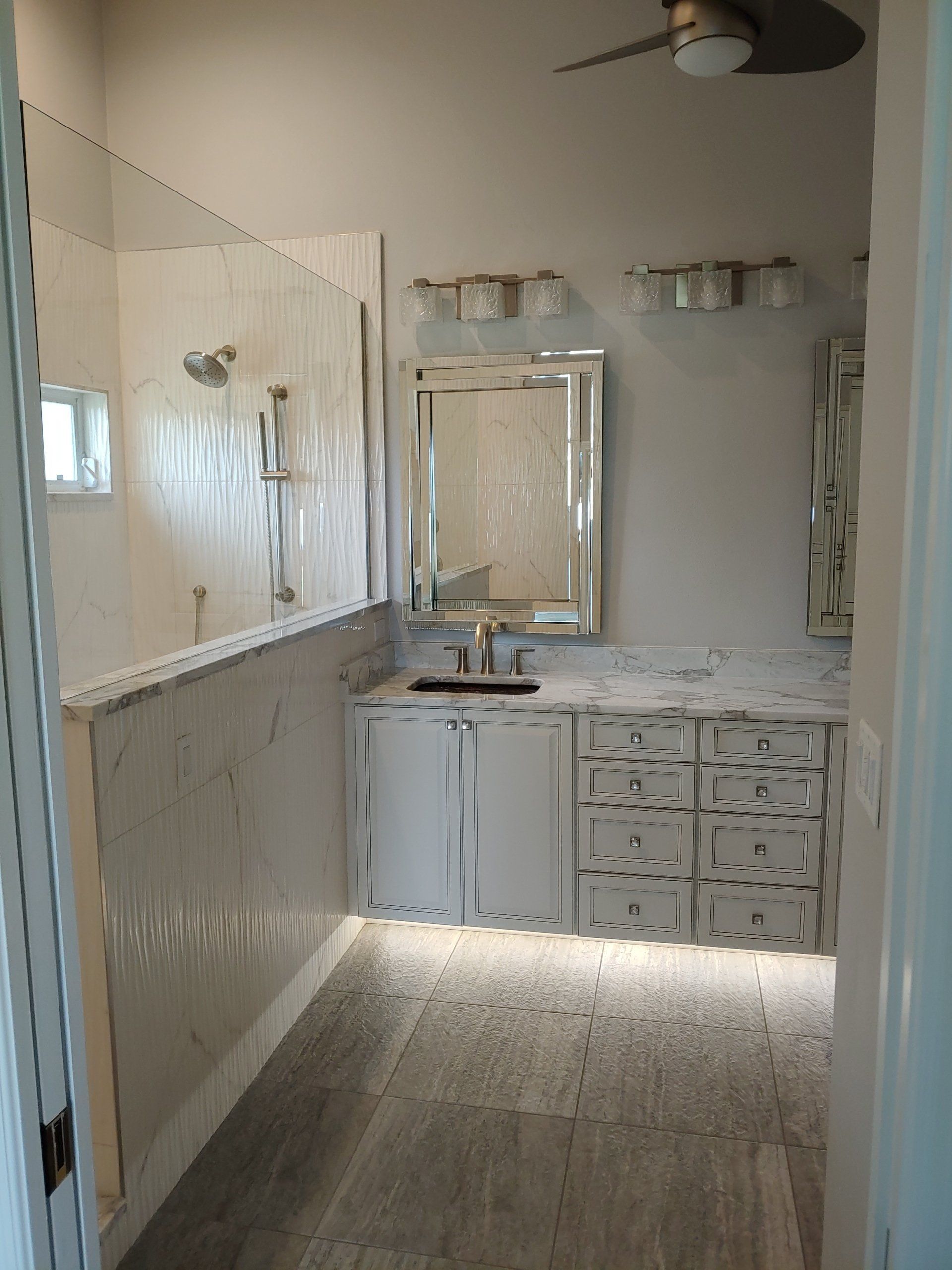 Bathroom with white textured walls, vanity, and shower. Gray floor. Mirror and two light fixtures above the vanity.