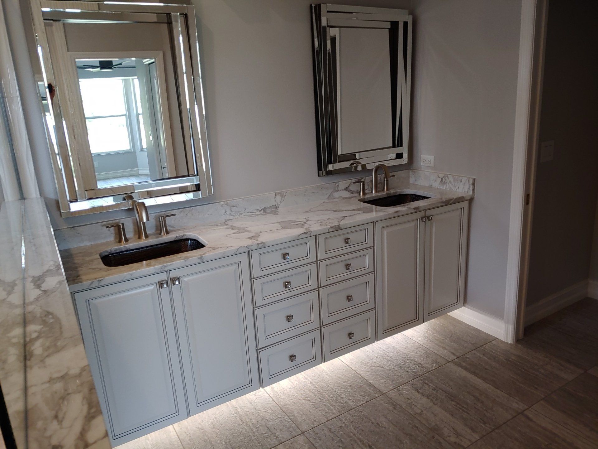 Bathroom vanity with two sinks, cabinets, marble countertop, and mirrors. Under-cabinet lighting illuminates the floor.