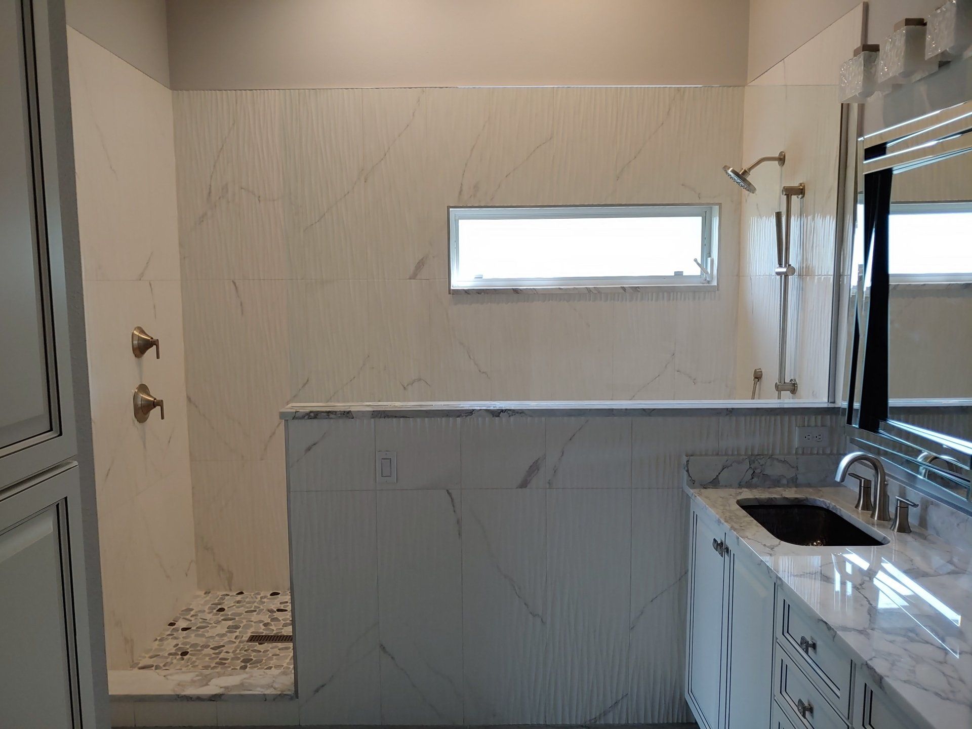 Bathroom with white marble shower and vanity, small window, and bronze fixtures.
