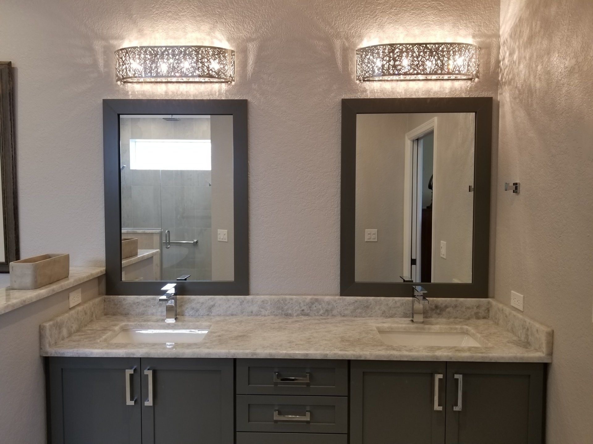 Bathroom with two mirrors, sinks, and gray cabinets, under patterned sconces.