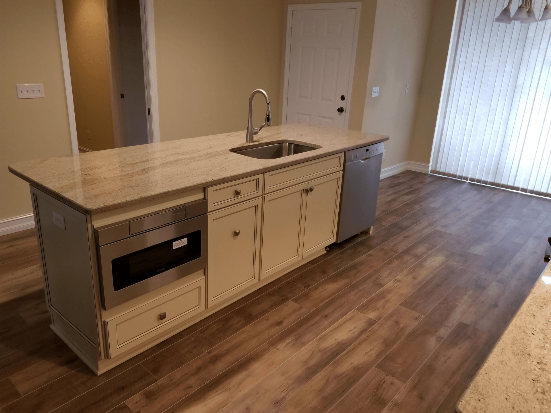 Kitchen island with a sink, dishwasher, and microwave on wood-look flooring, light-colored cabinets.