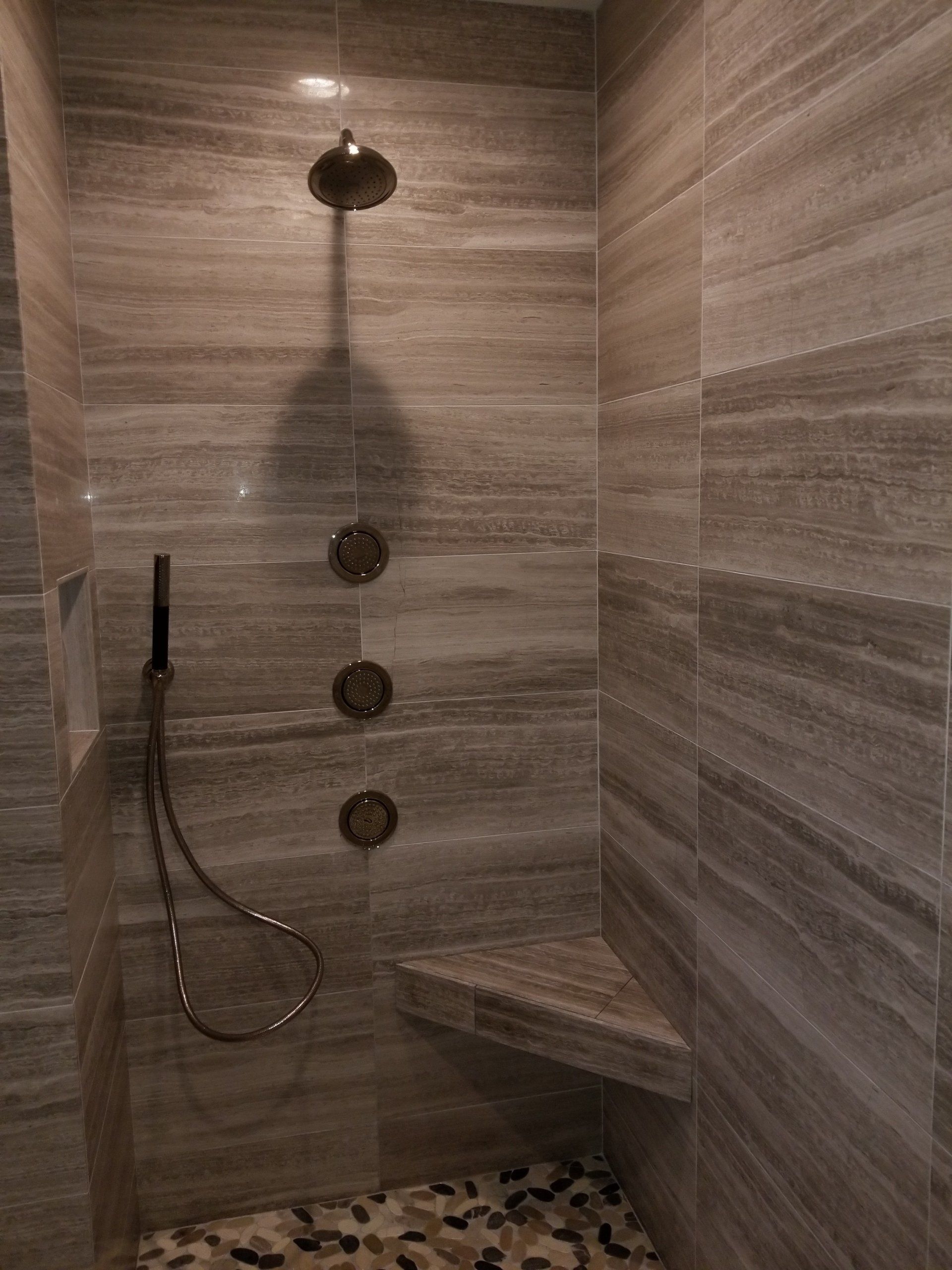 Shower with gray tile walls, pebble floor, shower head, and bench.