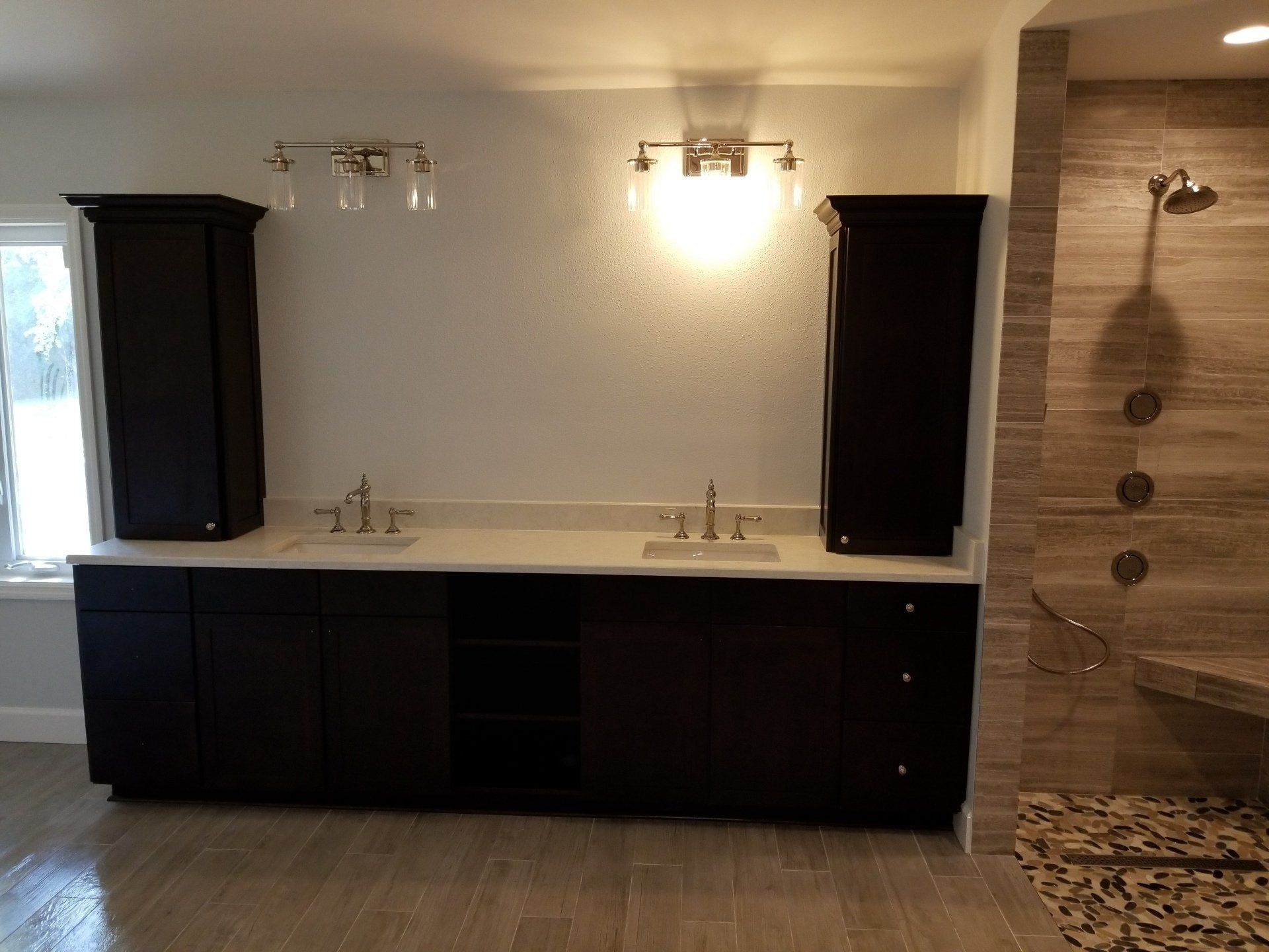 Bathroom with dark cabinets, double sinks, wall-mounted lights, and a tiled shower with pebbles on the floor.