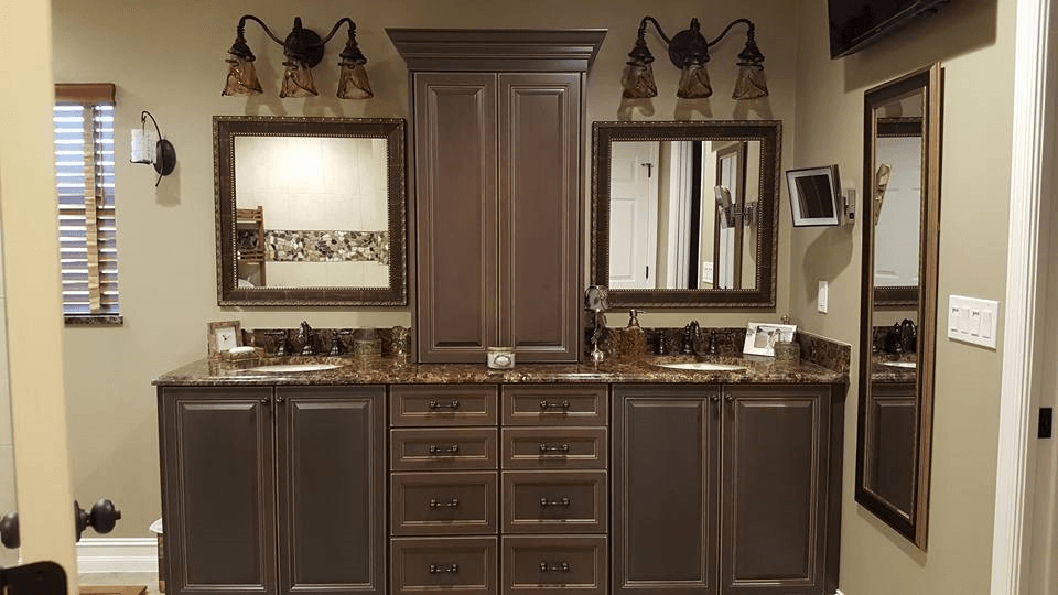 Bathroom with brown cabinets, mirrors, and lighting. The countertop is dark with a brown backsplash and beige walls.