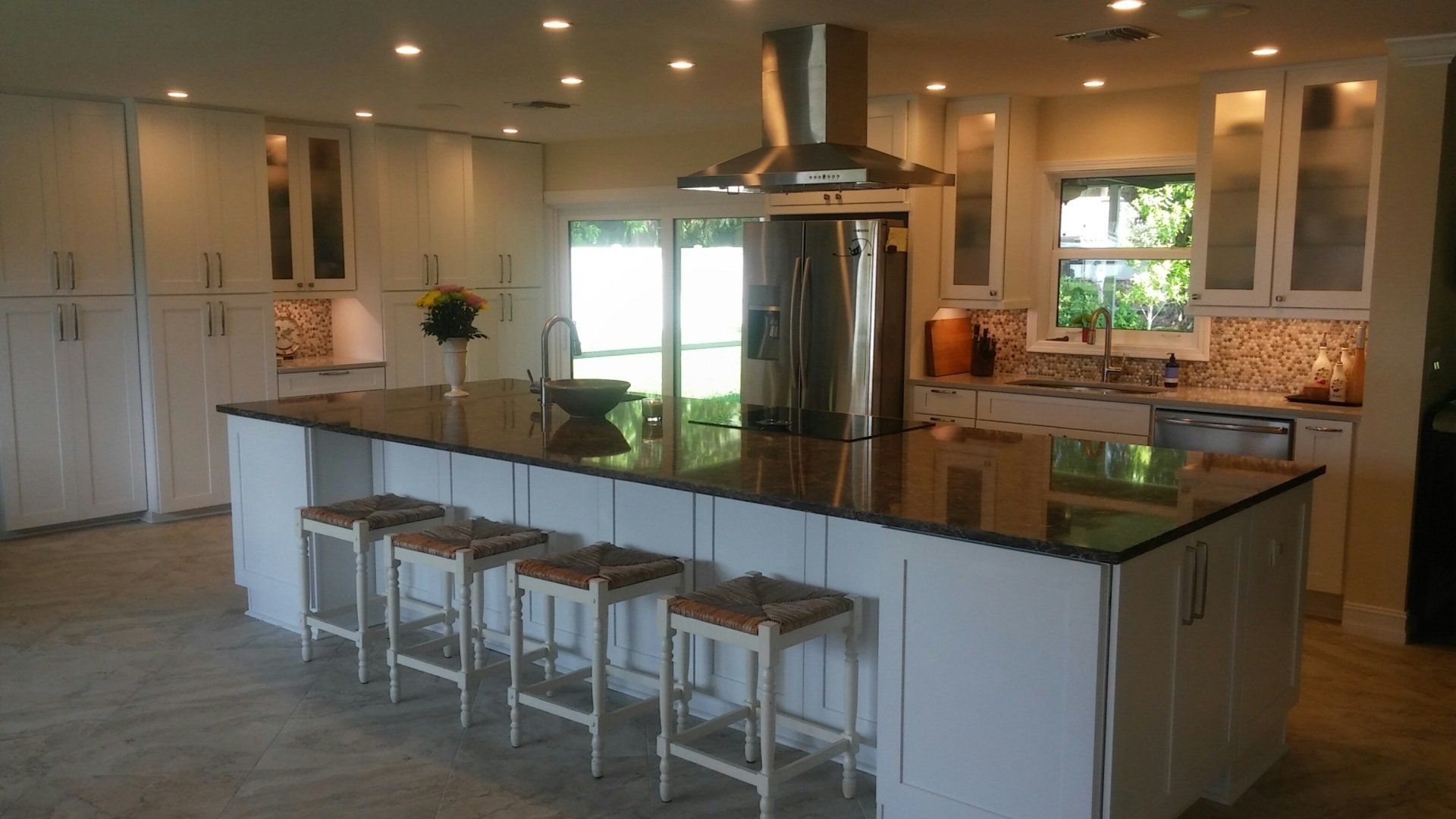 White kitchen with large island, dark countertop, stainless steel range hood, and stools.