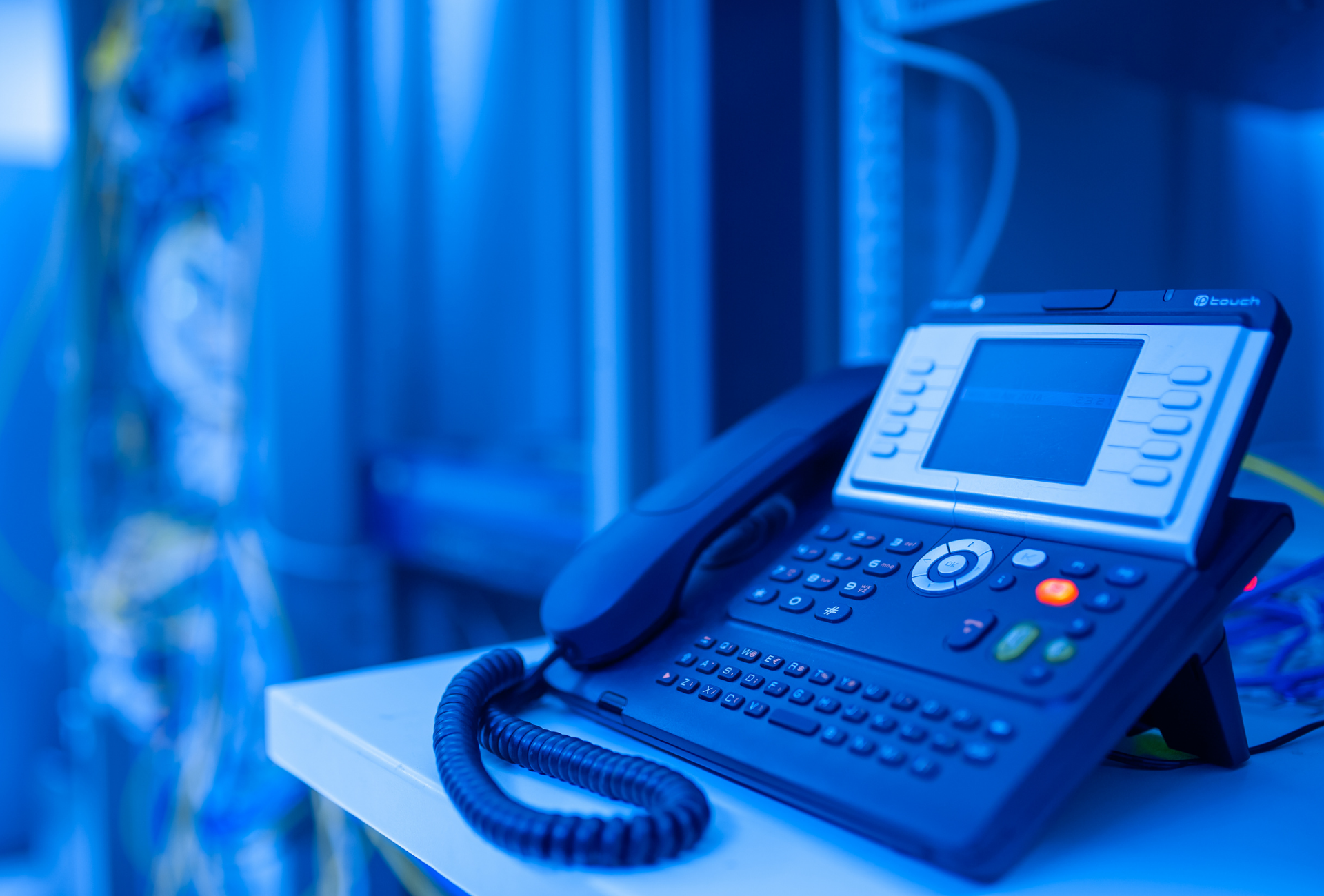 Blue-tinted image of a desktop IP phone on a white shelf in a server room, with network cables in the background.