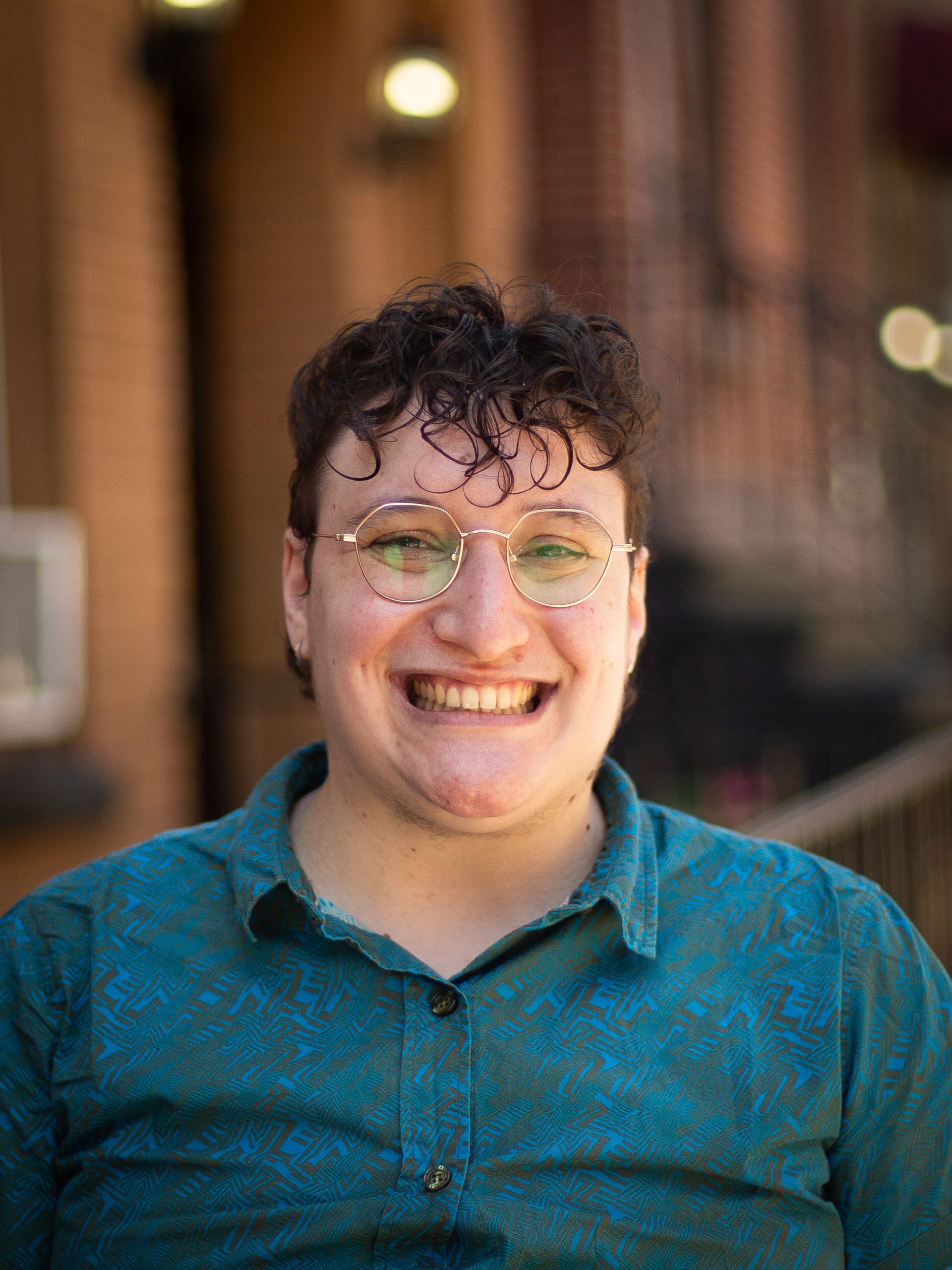 Smiling person in a teal shirt and glasses seated indoors with a blurred background