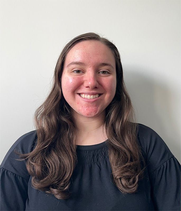 Woman with long, wavy brown hair smiles, wearing a black button-down shirt, near a window.