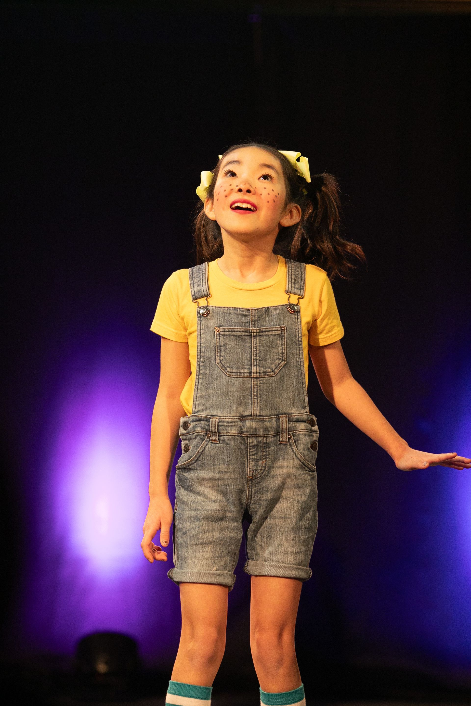 A little girl is standing on a stage wearing overalls and a yellow shirt.
