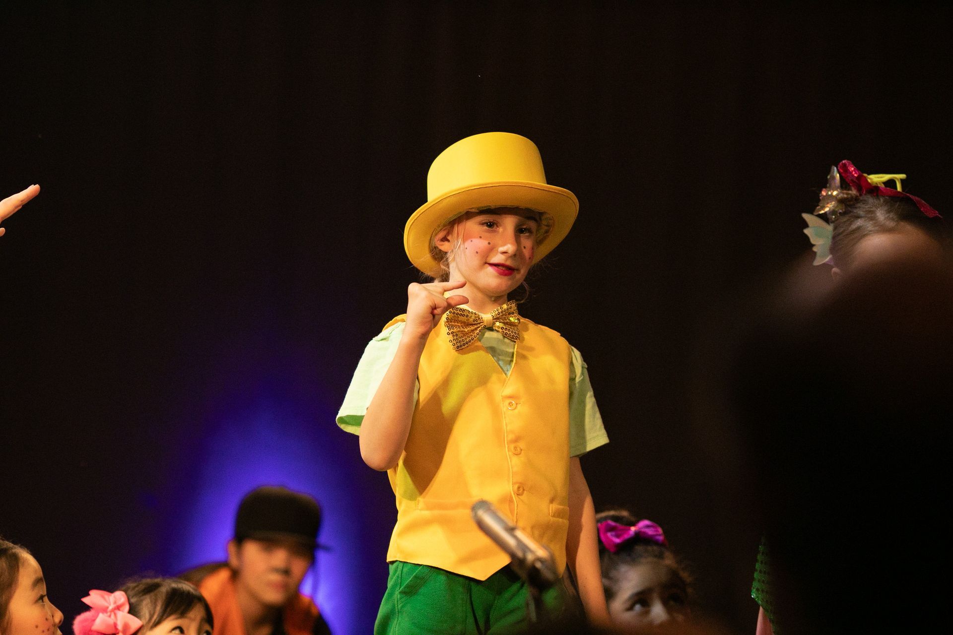 A young boy in a yellow hat and green vest is standing on a stage.