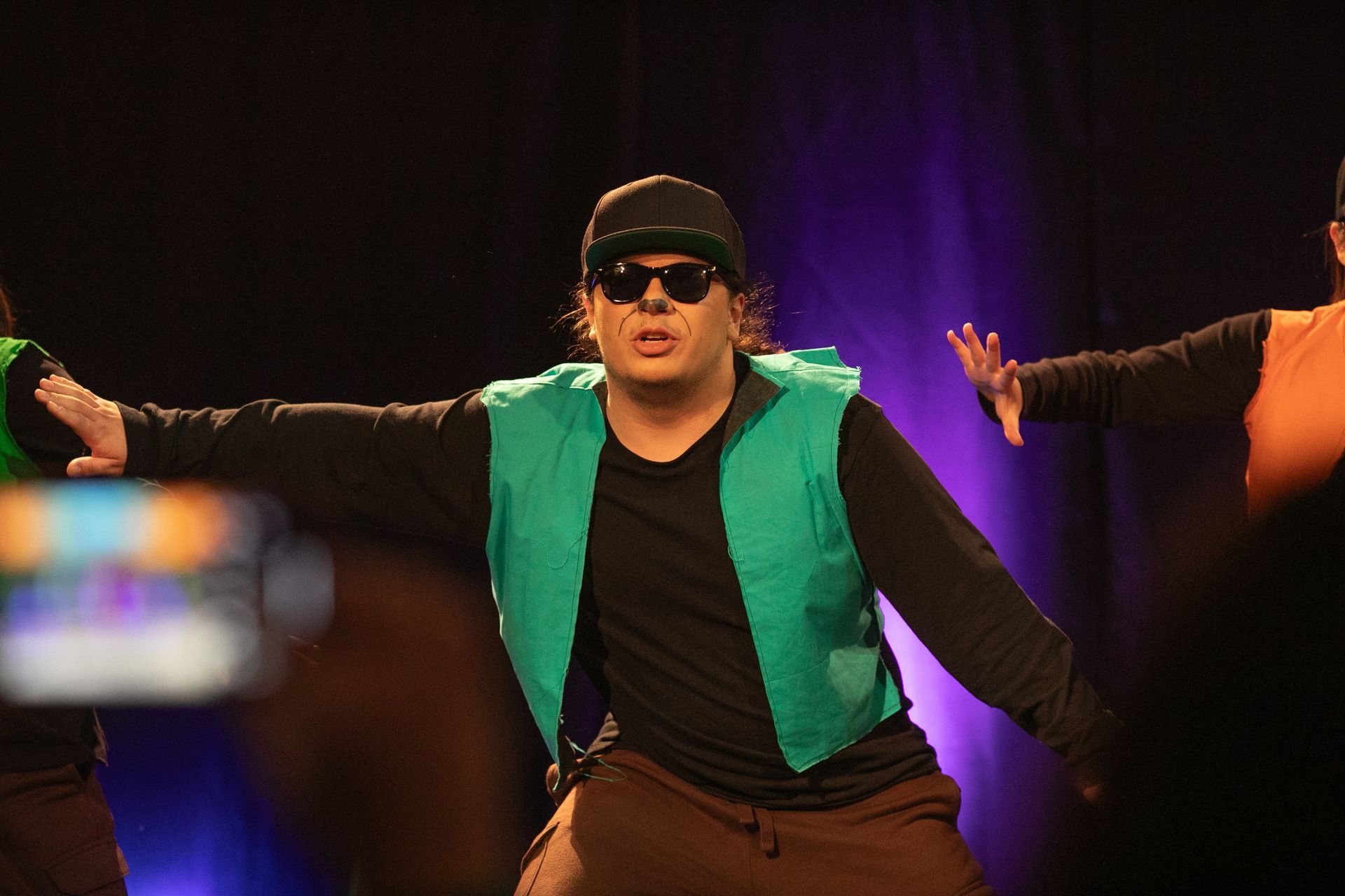 A man wearing sunglasses and a green vest is dancing on a stage.