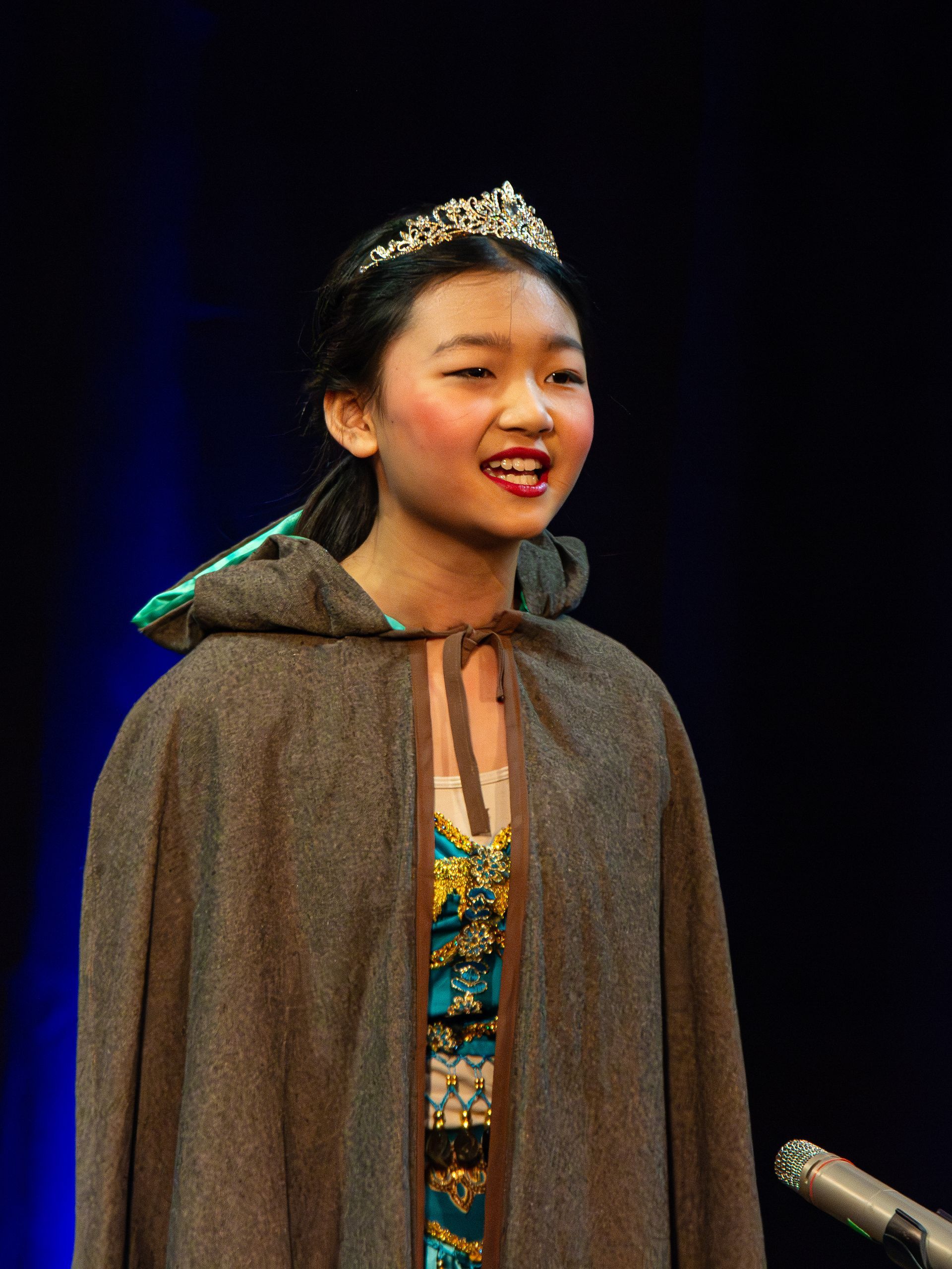 A young girl wearing a cape and tiara is singing into a microphone.