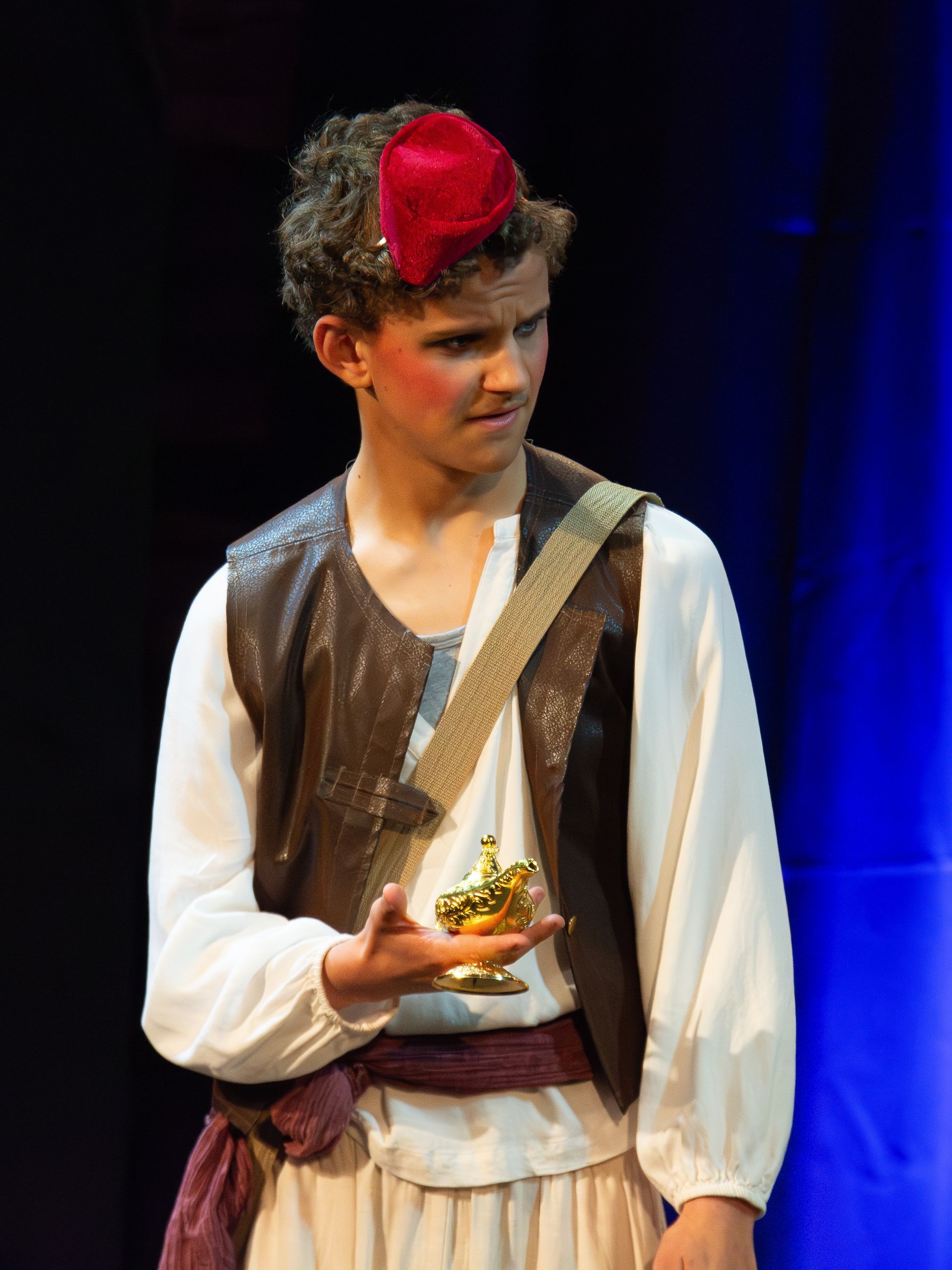 A young man in a costume is holding a lamp on a stage.